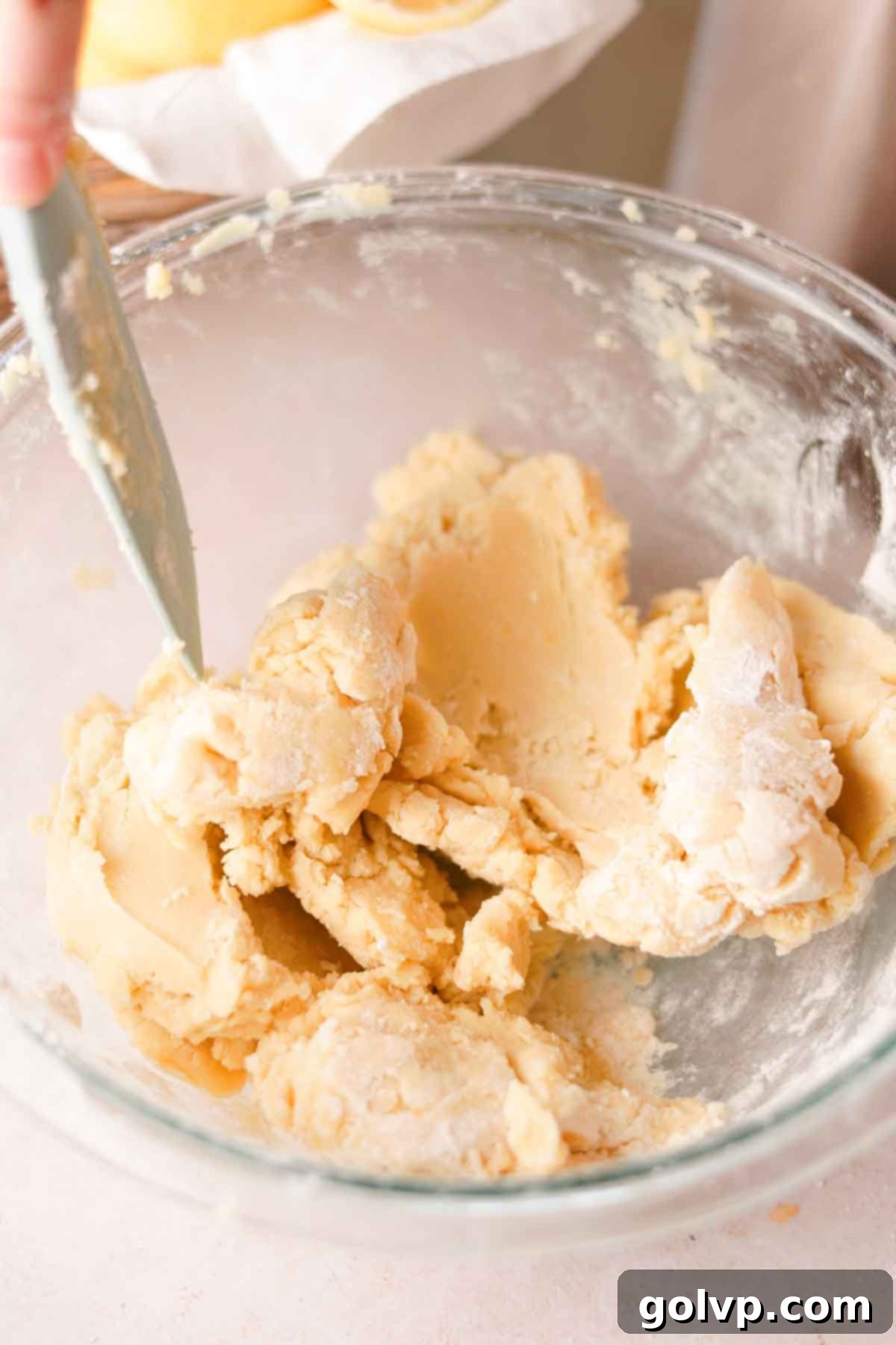 Folding all-purpose flour into a crumbly shortbread cookie dough mixture using a spatula in a large glass bowl.