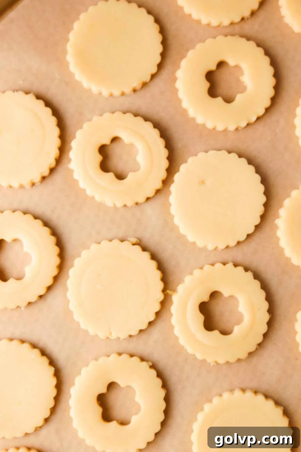 Unbaked shortbread cookies with scalloped edges and floral cut-outs in the middle arranged on a parchment-lined baking sheet.