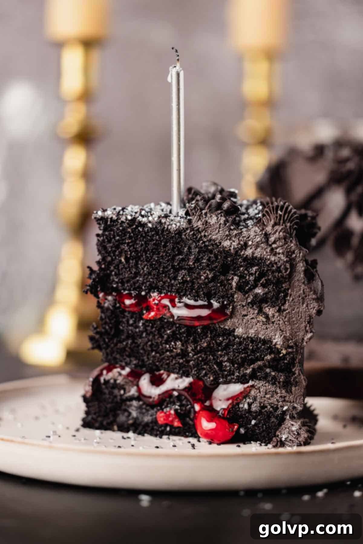 A perfect slice of black velvet cake, showing the contrasting black frosting and vivid red cherry filling, with a lit candle on top.
