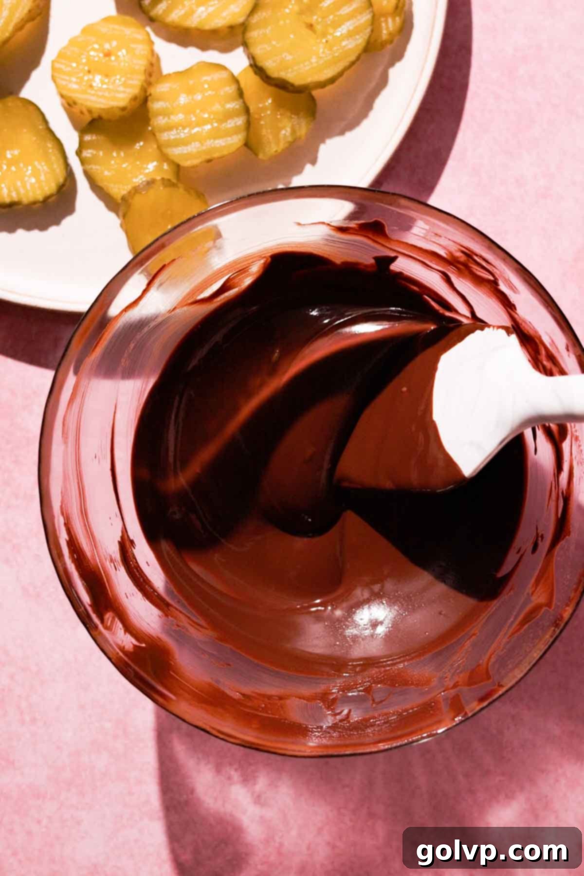 melted chocolate in glass bowl with rubber spatula and sliced pickles on the side