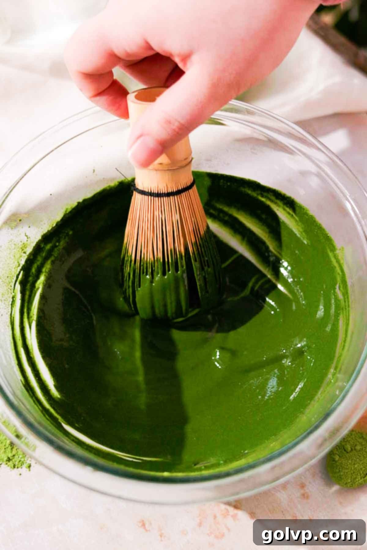 whisking vibrant green matcha and hot water in a large glass bowl with a traditional bamboo matcha whisk