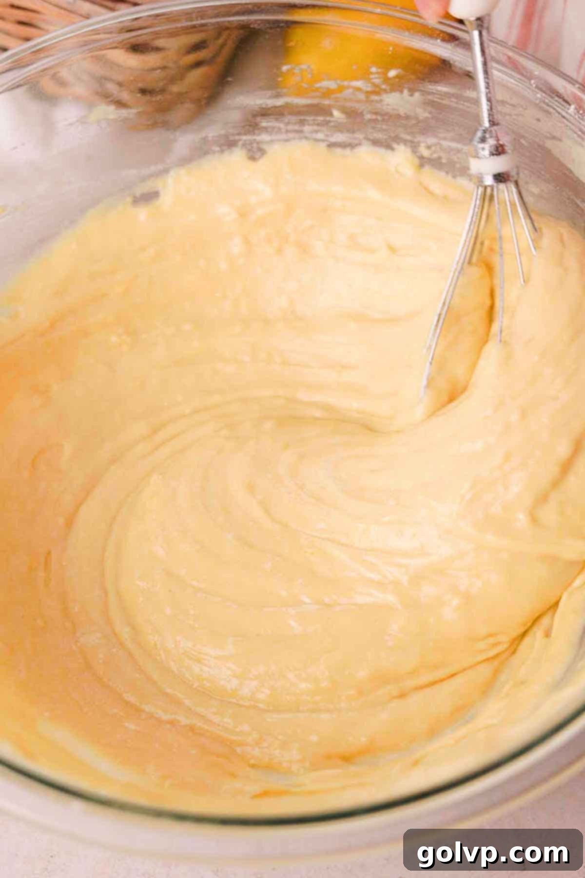 A whisk gently folding dry ingredients into the wet batter for lemon olive oil cake in a large glass bowl, being careful not to overmix.