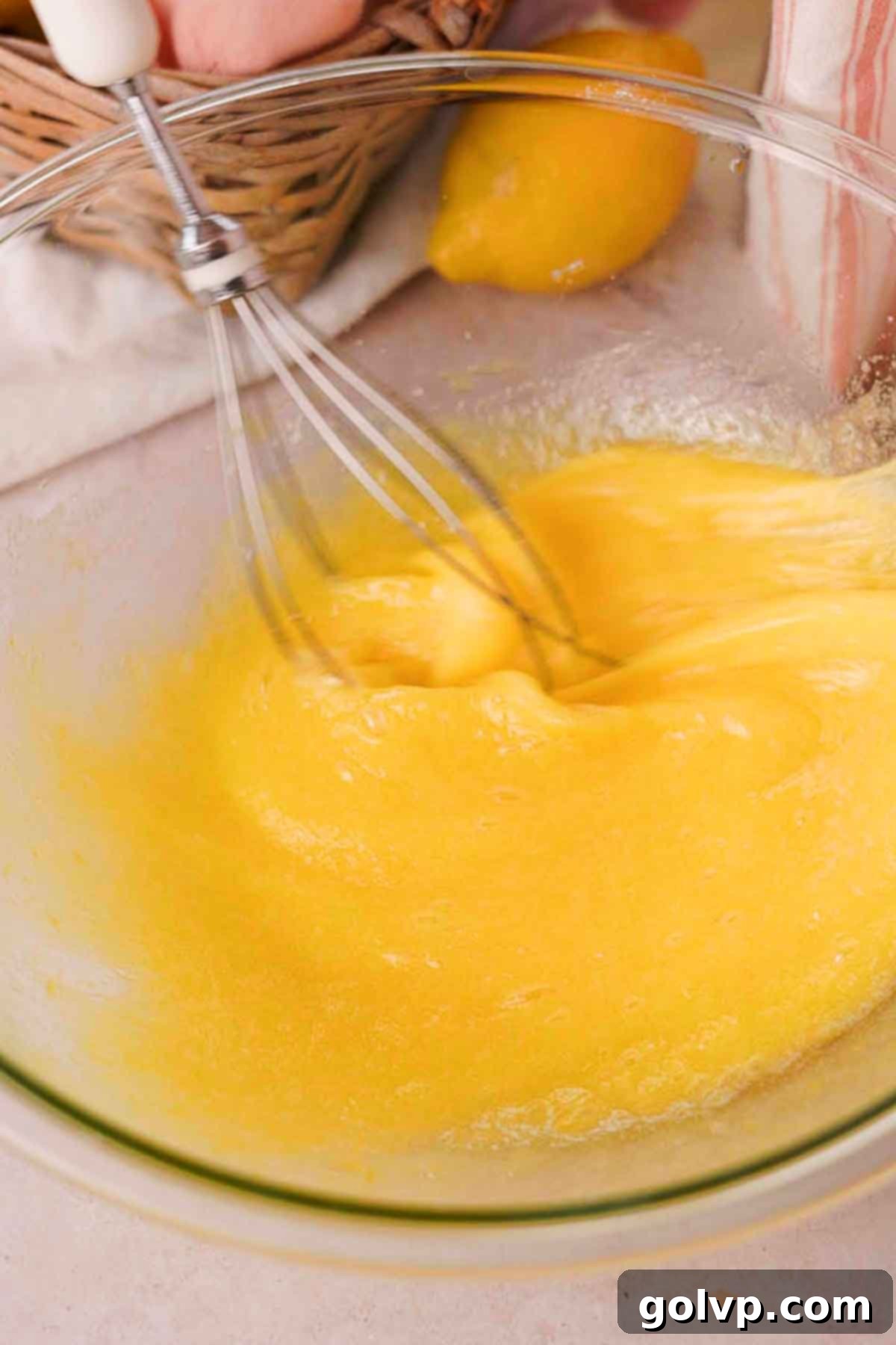 A wire whisk rapidly mixing lemon-infused sugar and eggs in a glass bowl, creating a frothy, pale yellow mixture.