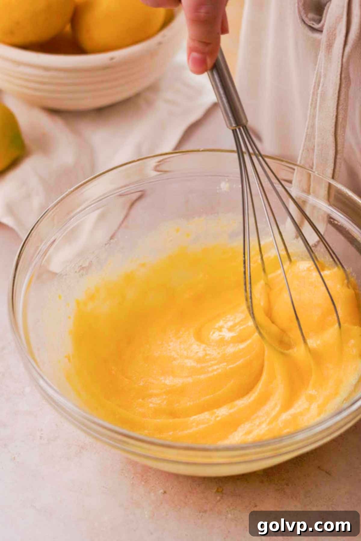 Whisking eggs, sugar, and lemon zest together in a glass bowl with a wire whisk, creating a thickened, pale mixture