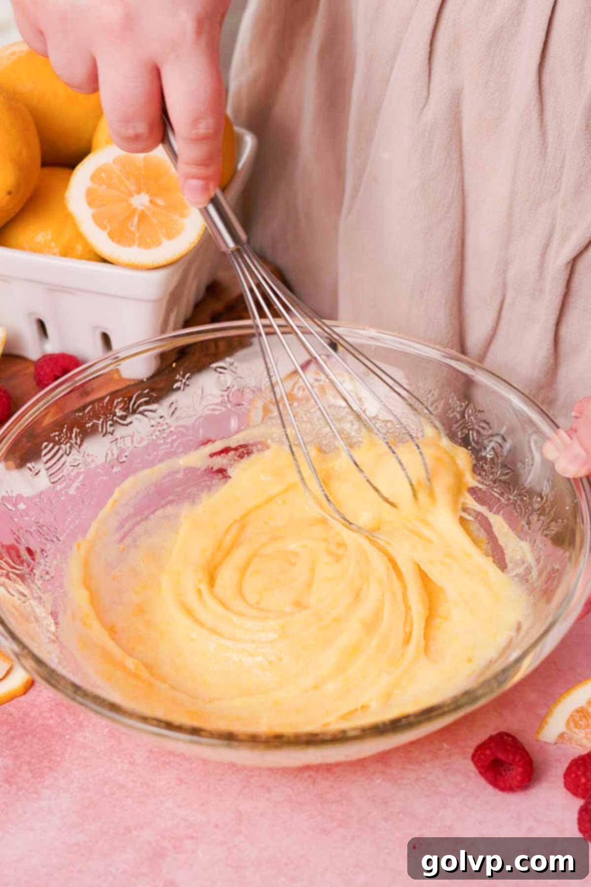 Whisking wet ingredients including egg, egg yolk, and lemon-infused sugar in a large glass bowl, creating a light and airy mixture