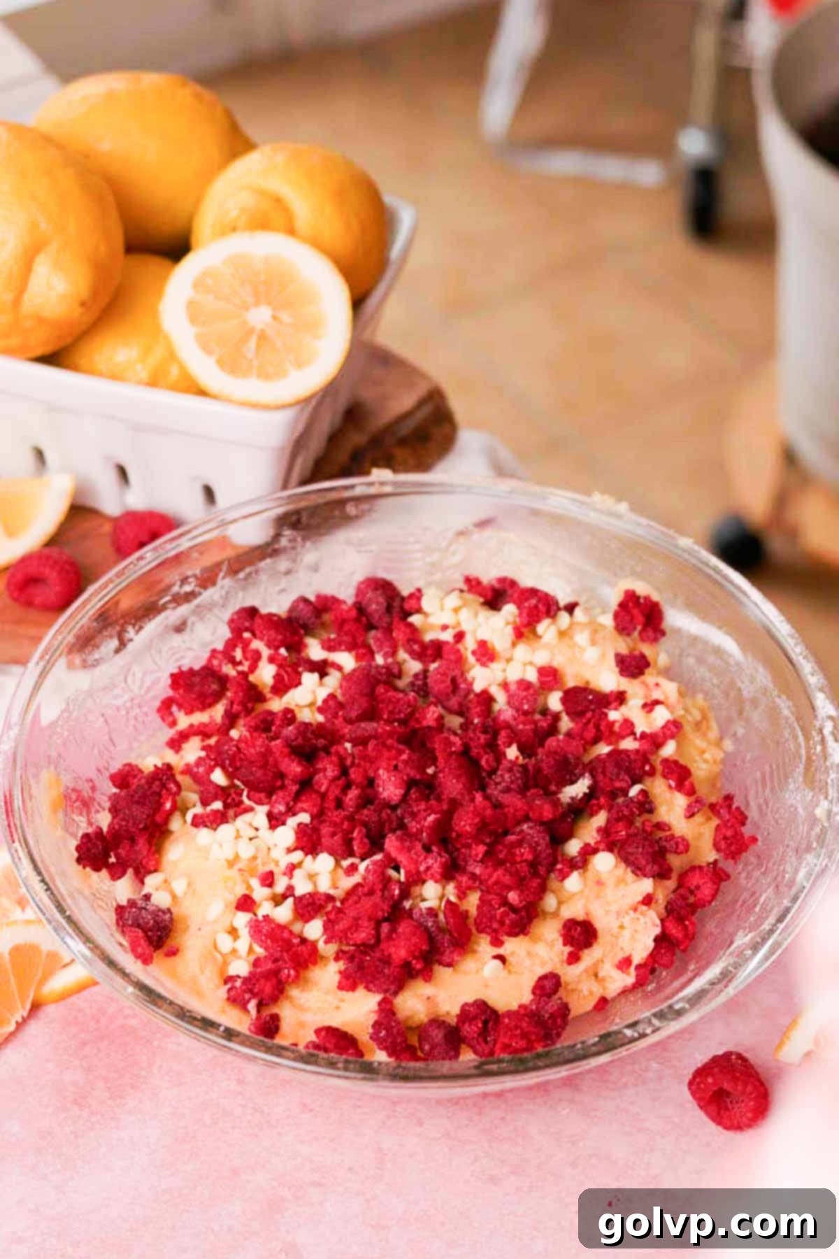 Adding broken up frozen raspberries and white chocolate chips into a large bowl of cookie dough for vibrant swirls and creamy pockets