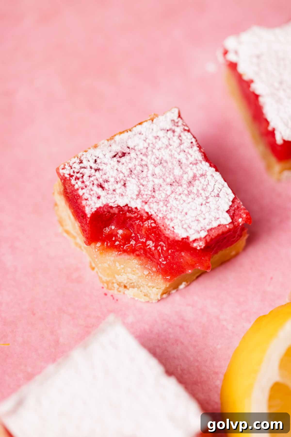 A single bitten lemon raspberry bar resting on pink countertop, revealing the vibrant filling and crisp crust.