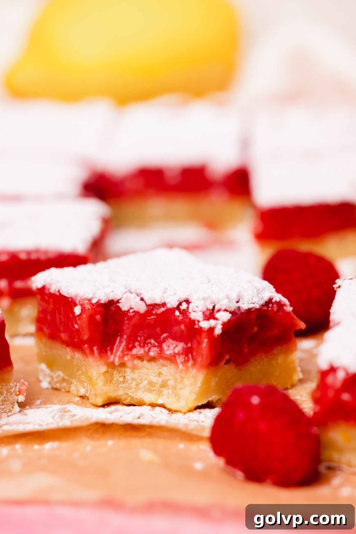 A perfectly cut lemon raspberry bar with a bite taken out, resting on parchment paper, surrounded by fresh raspberries.