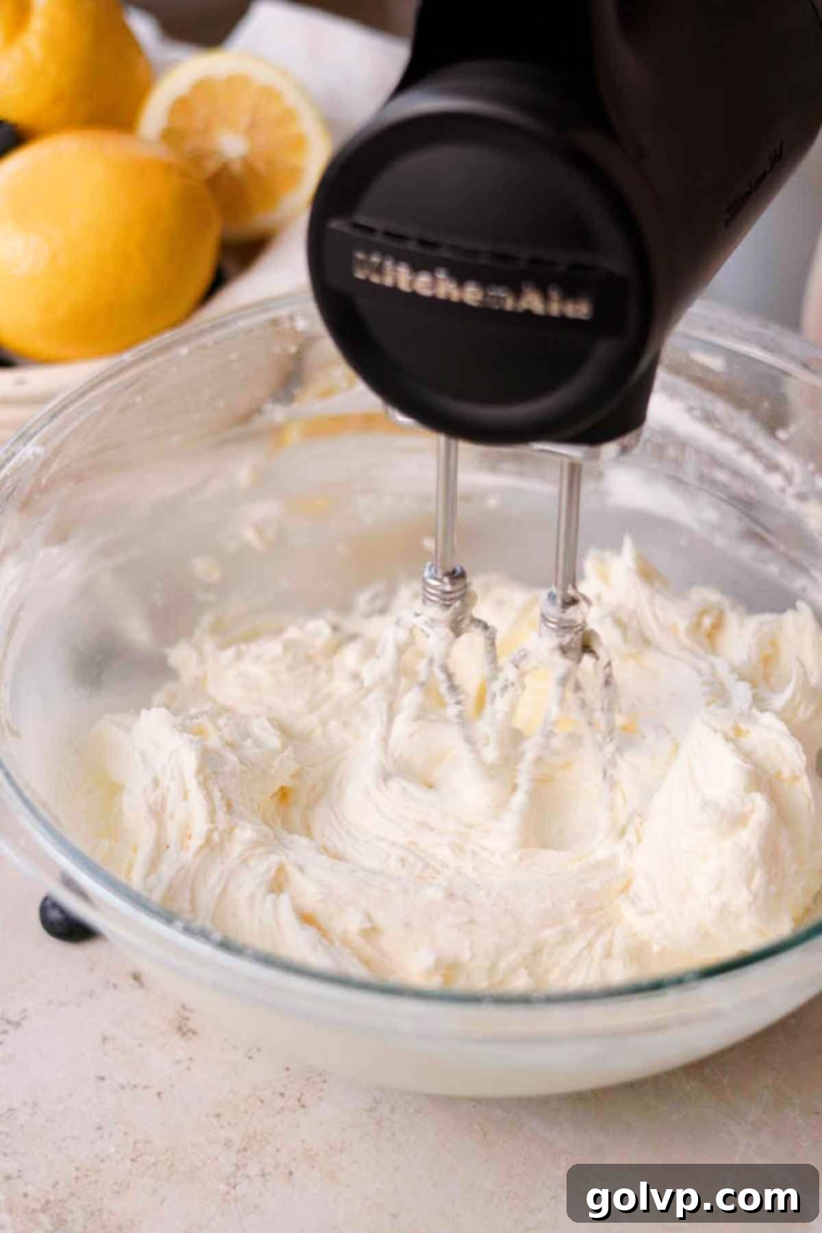 Whipping cream cheese frosting with an electric mixer in a large glass bowl, showing a smooth, thick consistency.