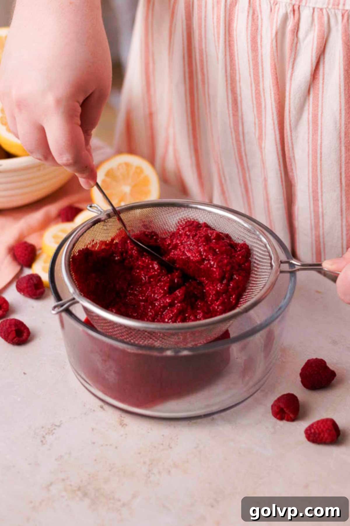 pushing mashed raspberries through a metal sieve with a metal spoon into a glass container