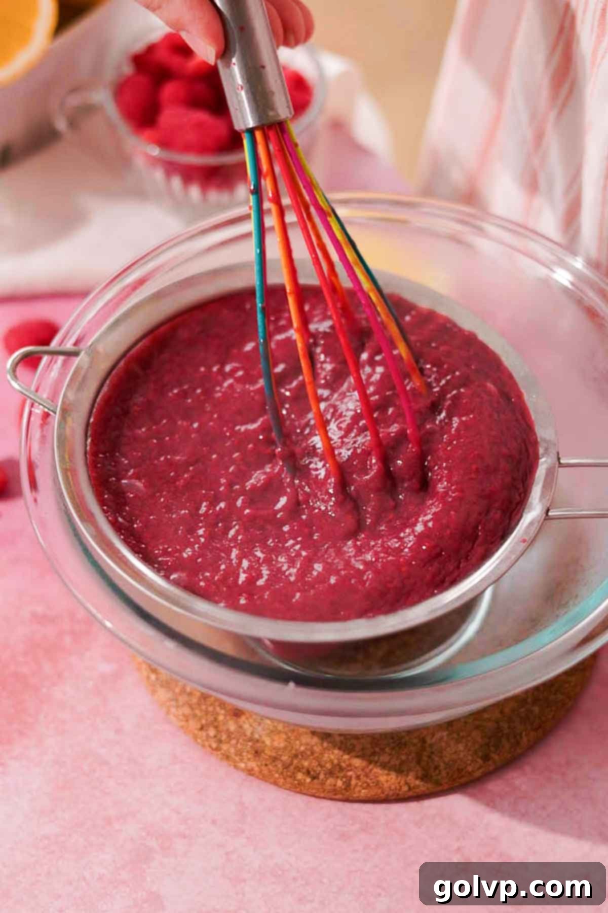 straining raspberry curd with silicone whisk through a metal sieve on top of a glass bowl