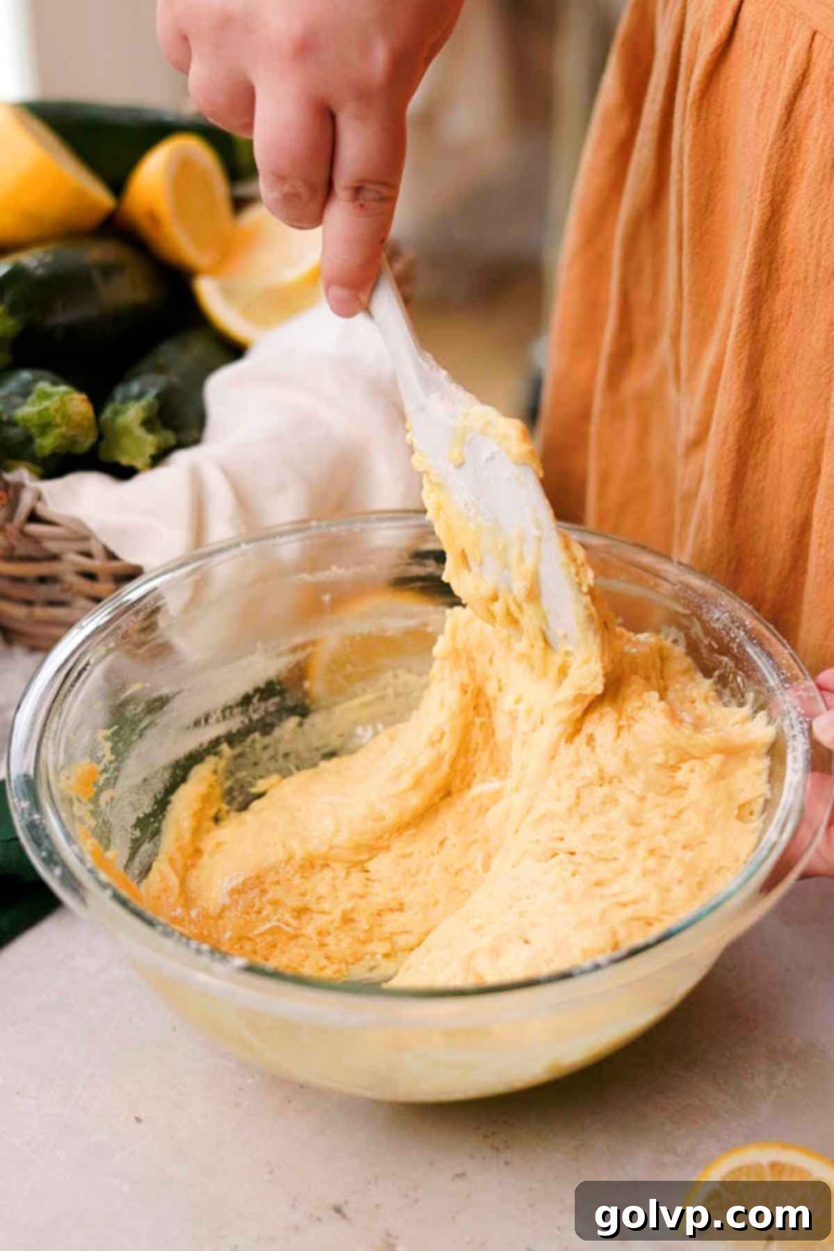 A silicone spatula gently folding sifted flour, baking powder, and baking soda into the wet lemon zucchini bread batter in a large bowl.