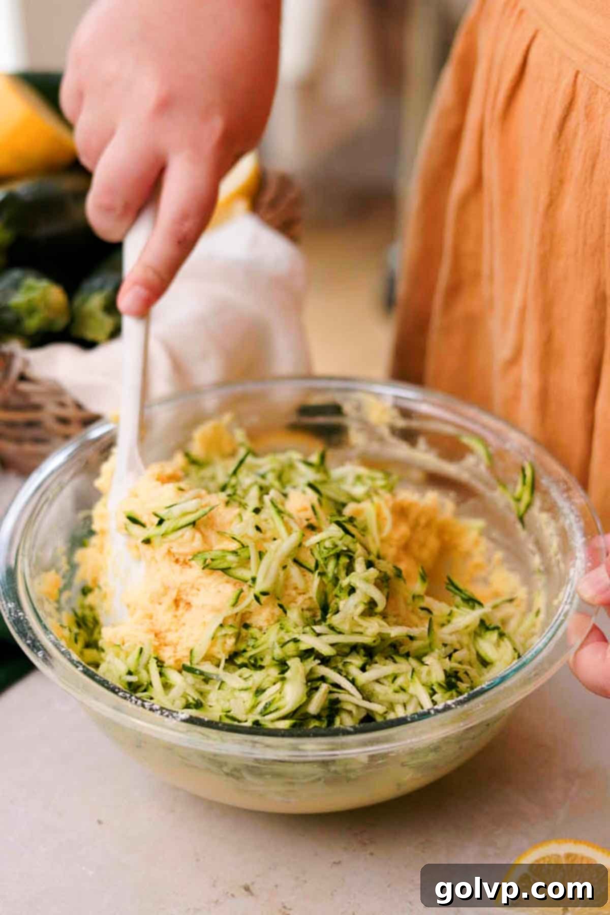 Hands carefully folding shredded zucchini into the lemon bread batter using a silicone spatula, ensuring even distribution.