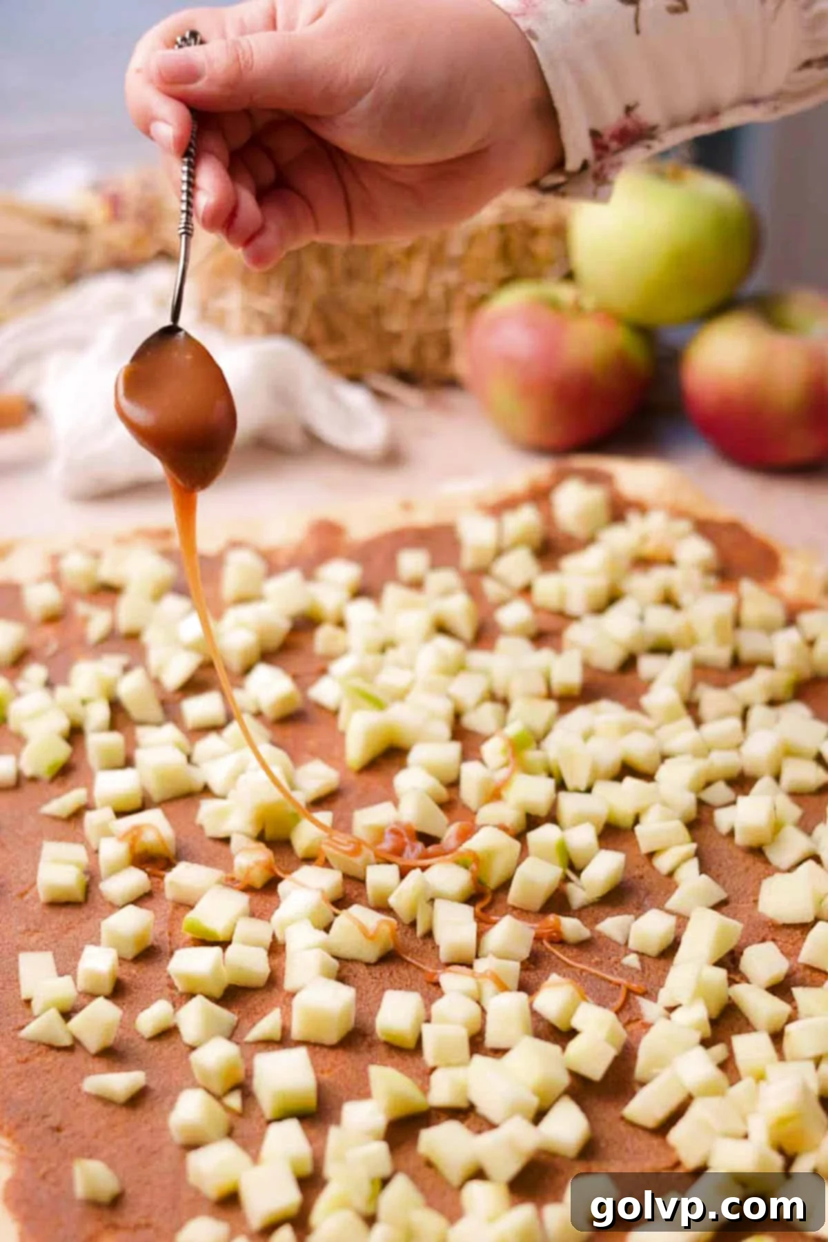 Carefully drizzling rich salted caramel sauce over the spread cinnamon sugar butter and chopped apples on the rolled-out cinnamon roll dough.
