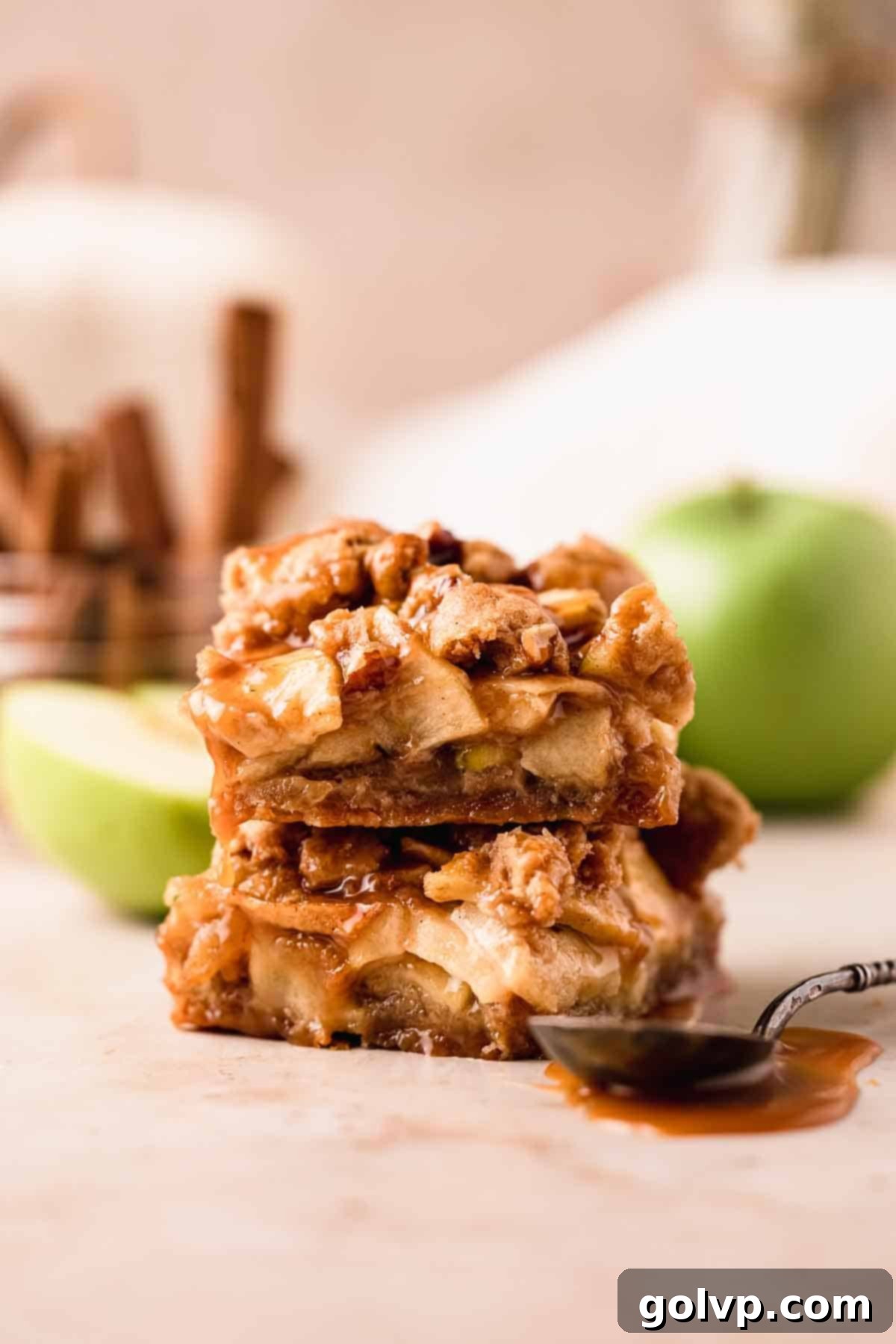 A stack of golden-brown salted caramel apple pie bars, with a spoon nearby, showcasing their inviting caramel drizzle and delicious crumbly topping.