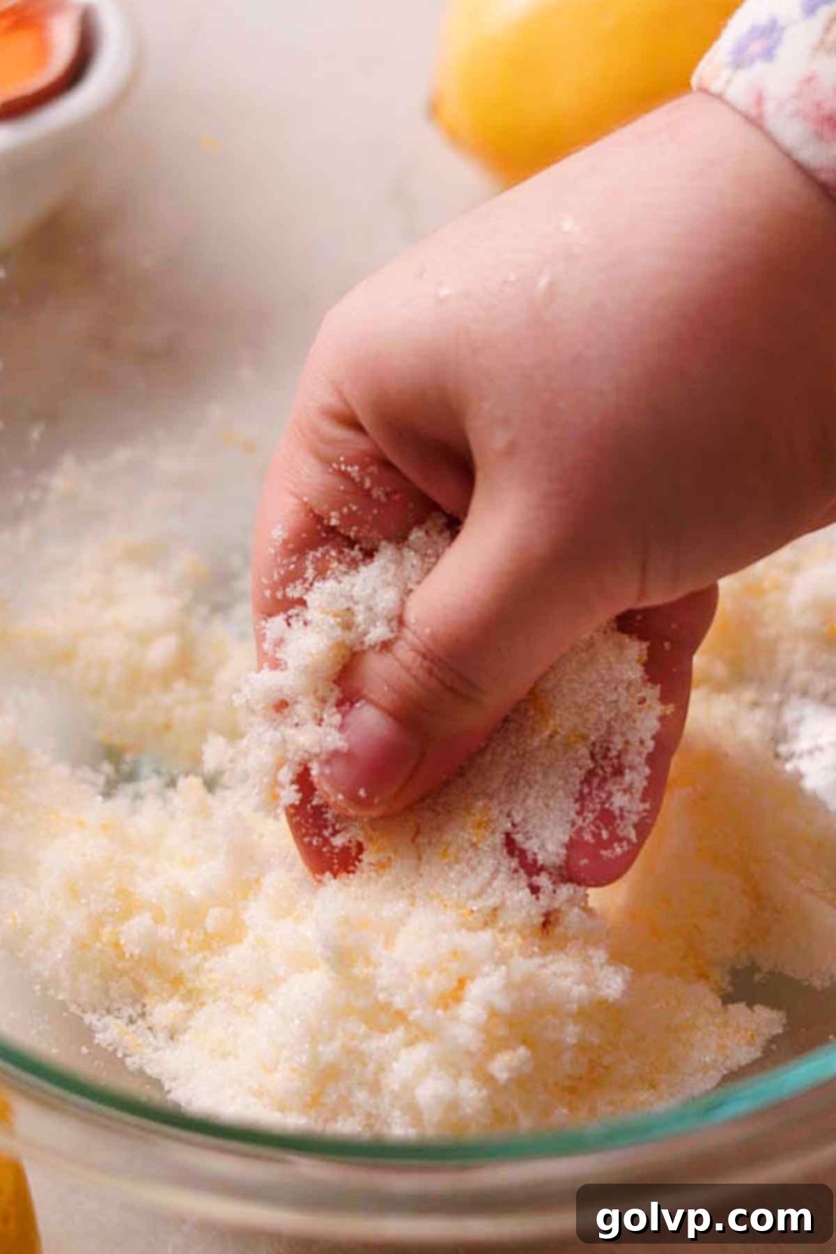 A close-up of hands rubbing lemon zest and granulated sugar together in a bowl, infusing the sugar with lemon oils