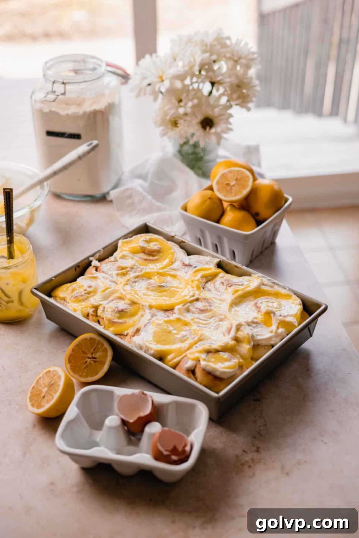 Arrangement of lemon rolls on a countertop with fresh lemons and flowers, highlighting their appealing presentation.