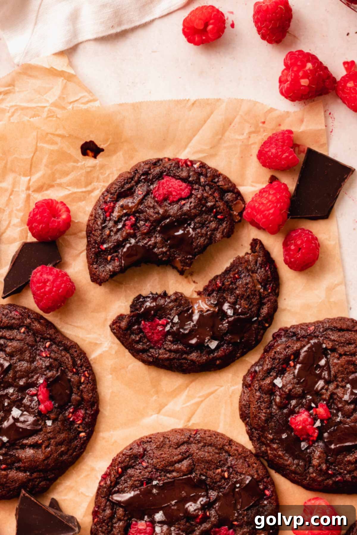 chocolate raspberry cookie broken in half on brown parchment paper with fresh raspberries around