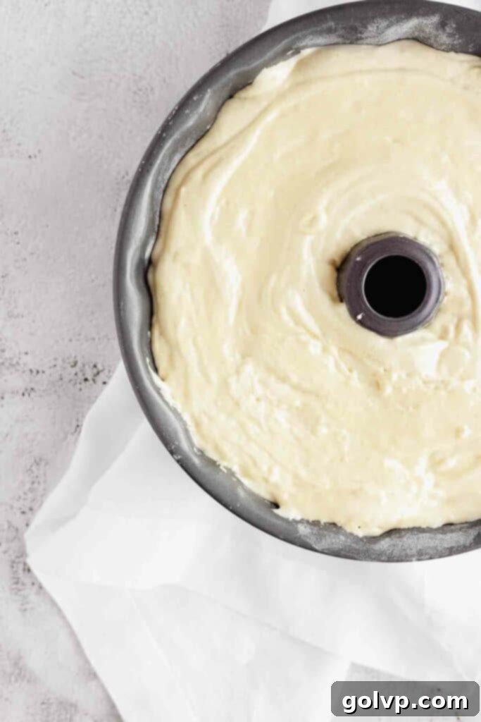 A spatula smoothing the light, airy Sharlotka batter into a greased and floured Bundt pan, covering the apple slices.