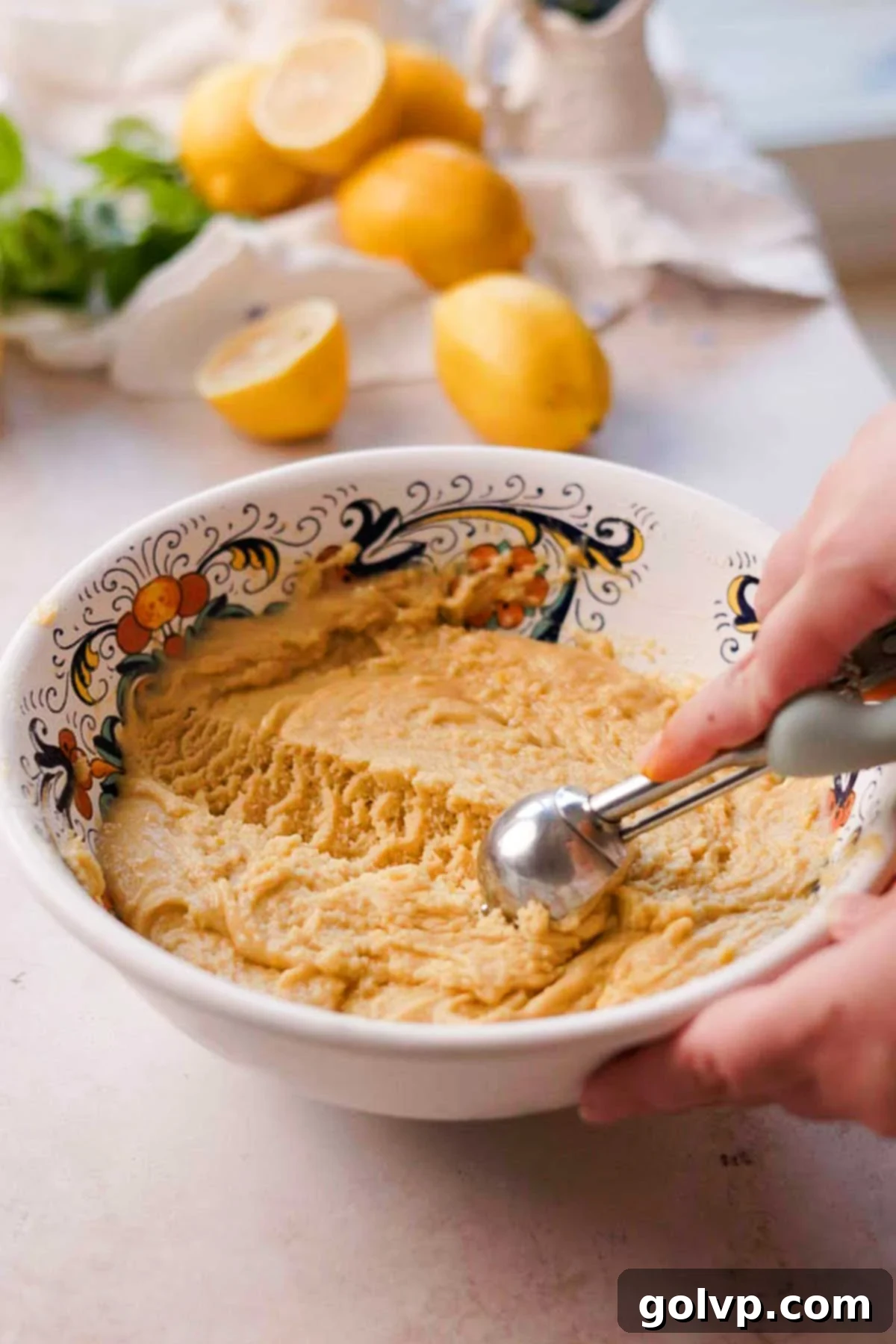 A metal scoop portions out the lemon olive oil cookie dough onto a parchment-lined baking sheet.