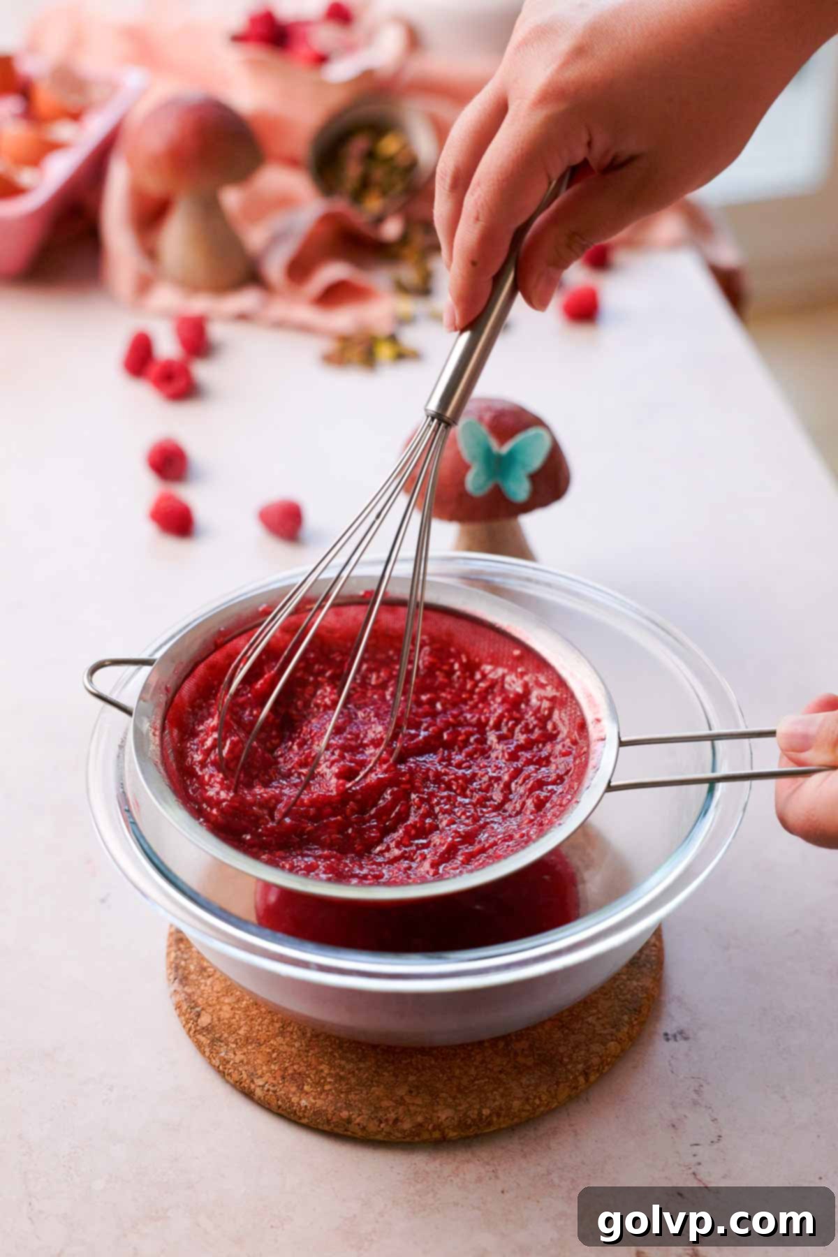 straining raspberry seeds through sieve with a wire whisk into a glass bowl