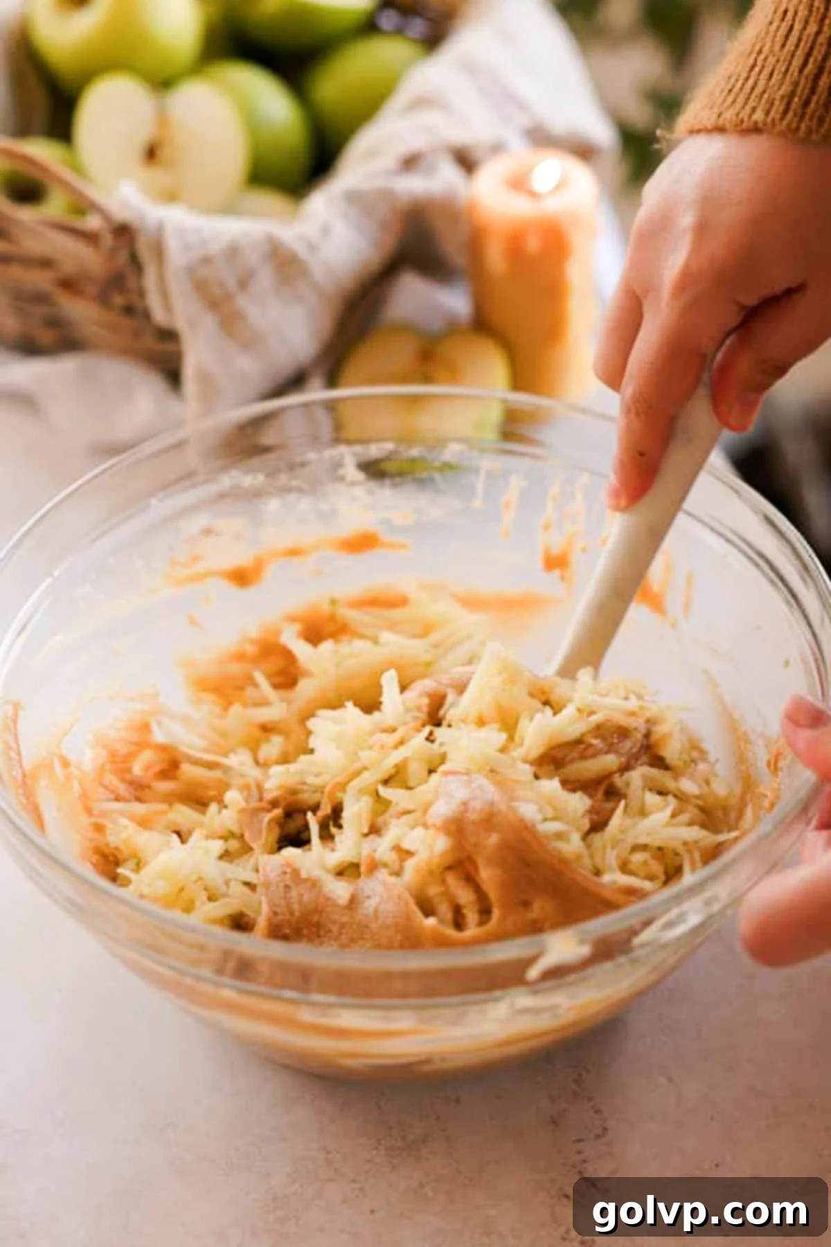 Folding shredded apples into muffin batter in a large glass bowl with a spatula