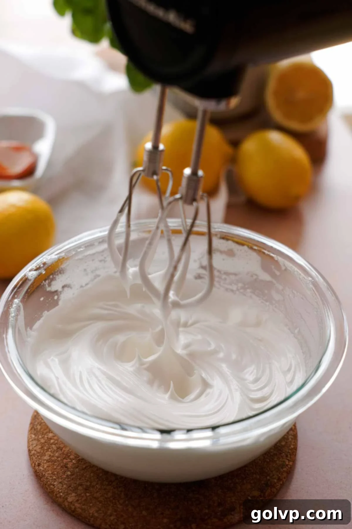 Whipping glossy Swiss meringue to stiff peaks with an electric hand mixer in a glass bowl.