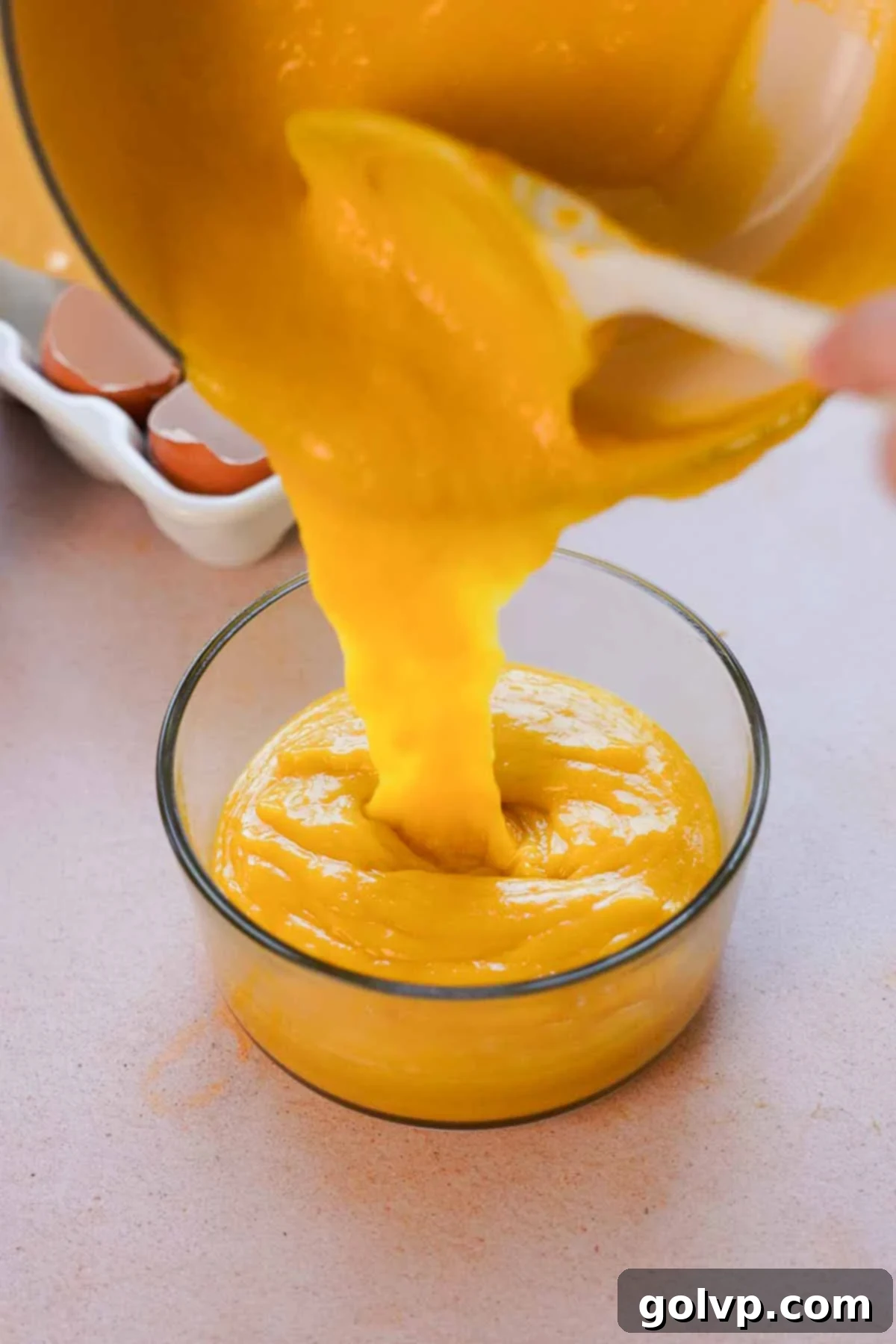Warm mango curd being poured from a saucepan into a clear glass container, prepared for chilling in the refrigerator.