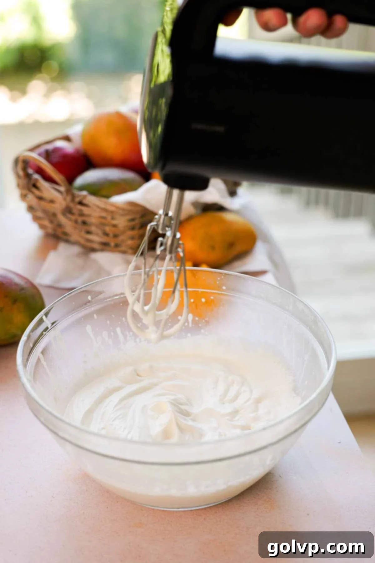 Heavy whipping cream being whipped to soft peaks in a large glass bowl using an electric mixer, indicating the first step for the tiramisu filling.