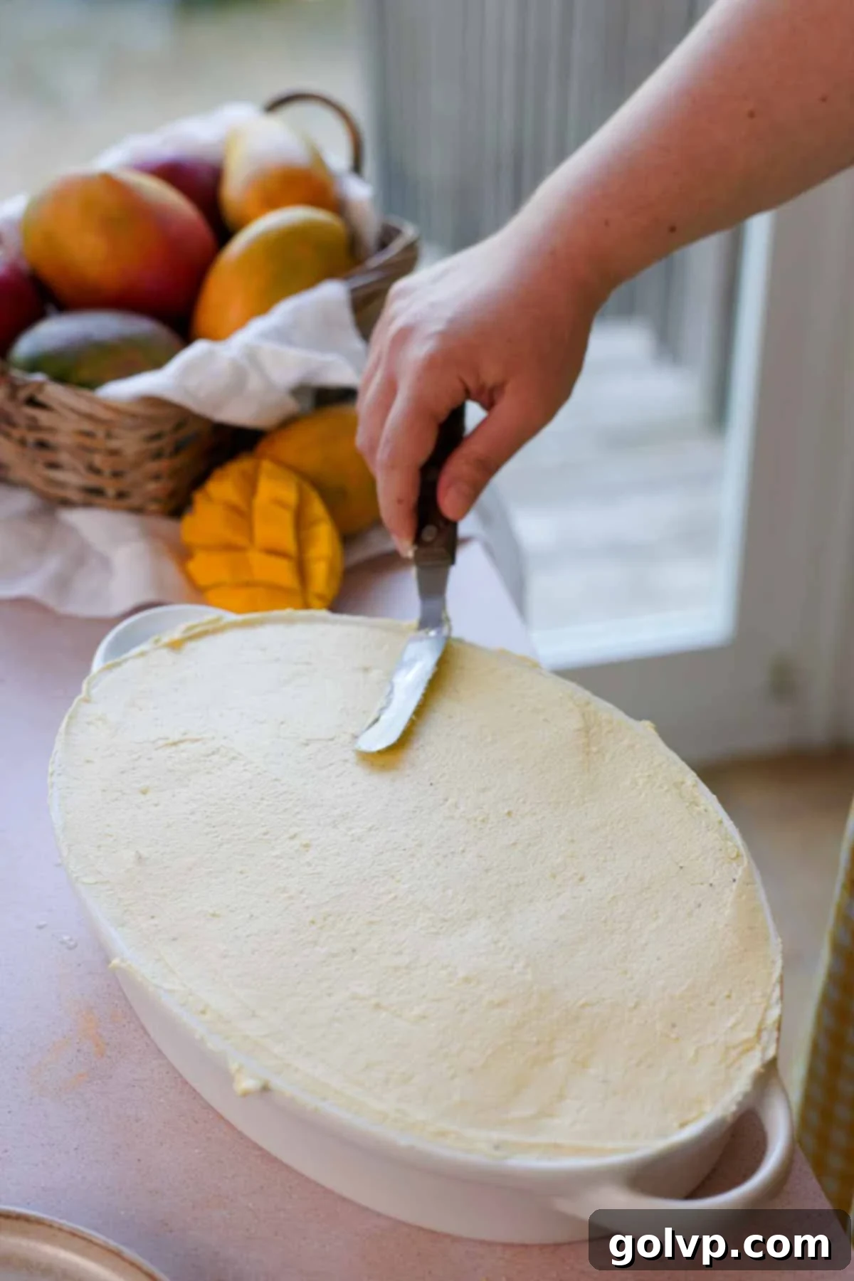 A spatula smoothing the top layer of the mango tiramisu filling in a baking dish, preparing it for chilling and final decoration.