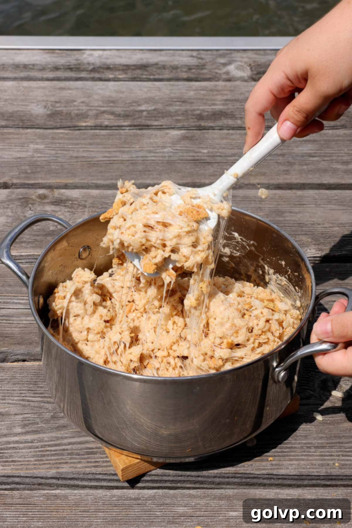 A pot filled with melted marshmallow and butter mixture, being combined with Rice Krispies cereal and graham cracker pieces using a spatula.