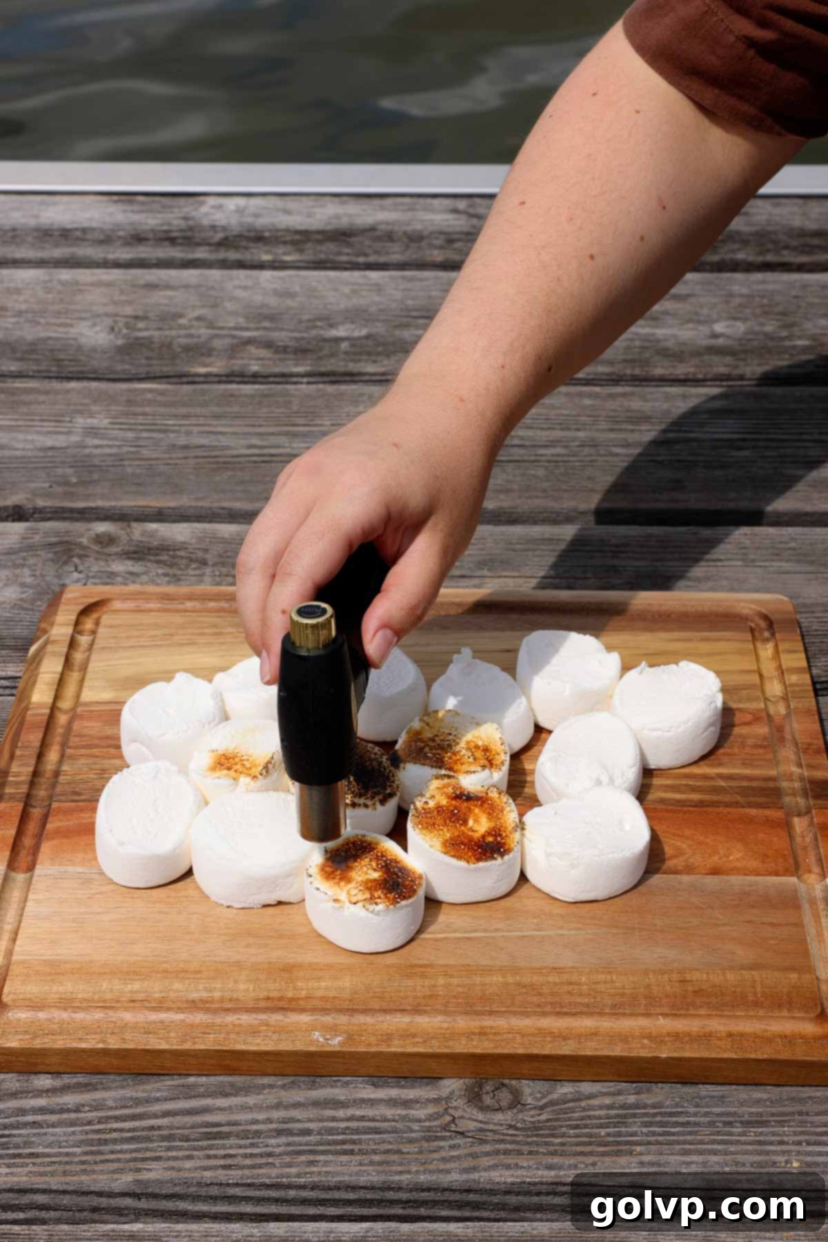 A kitchen torch being used to char and toast large marshmallows on a wooden cutting board, creating a slightly blackened exterior.