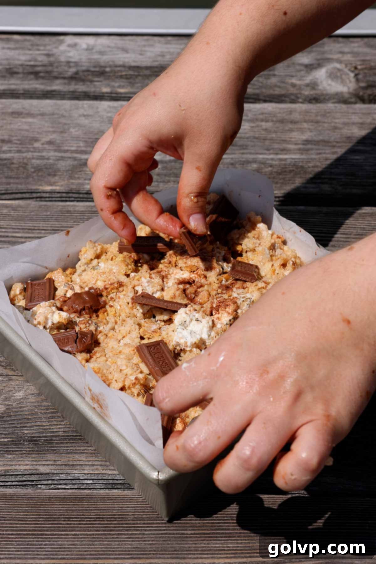 Small pieces of milk chocolate being placed onto the surface of S'mores Rice Krispies Treats in a parchment-lined pan.