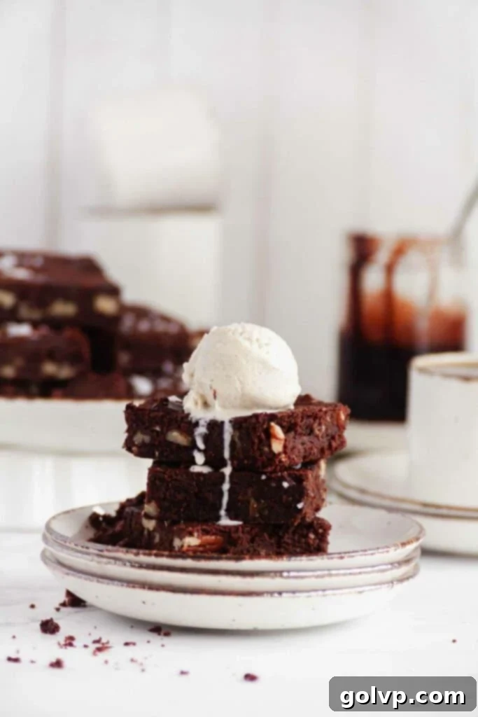 Stack of four chewy salted pecan brownies, topped with a scoop of vanilla ice cream and chocolate drizzle, set against a dark background.