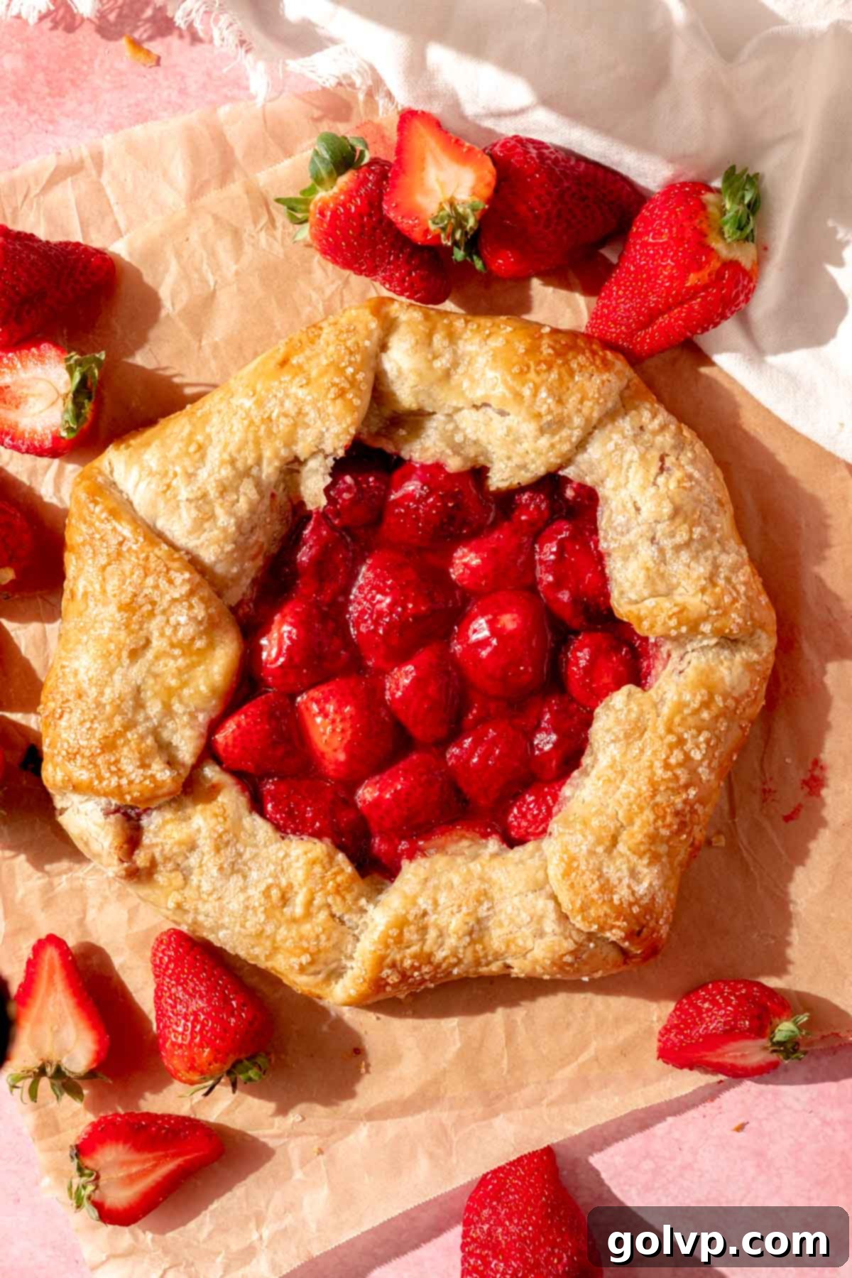 A beautifully rustic strawberry galette with a gooey strawberry filling, surrounded by fresh strawberries on a light background.
