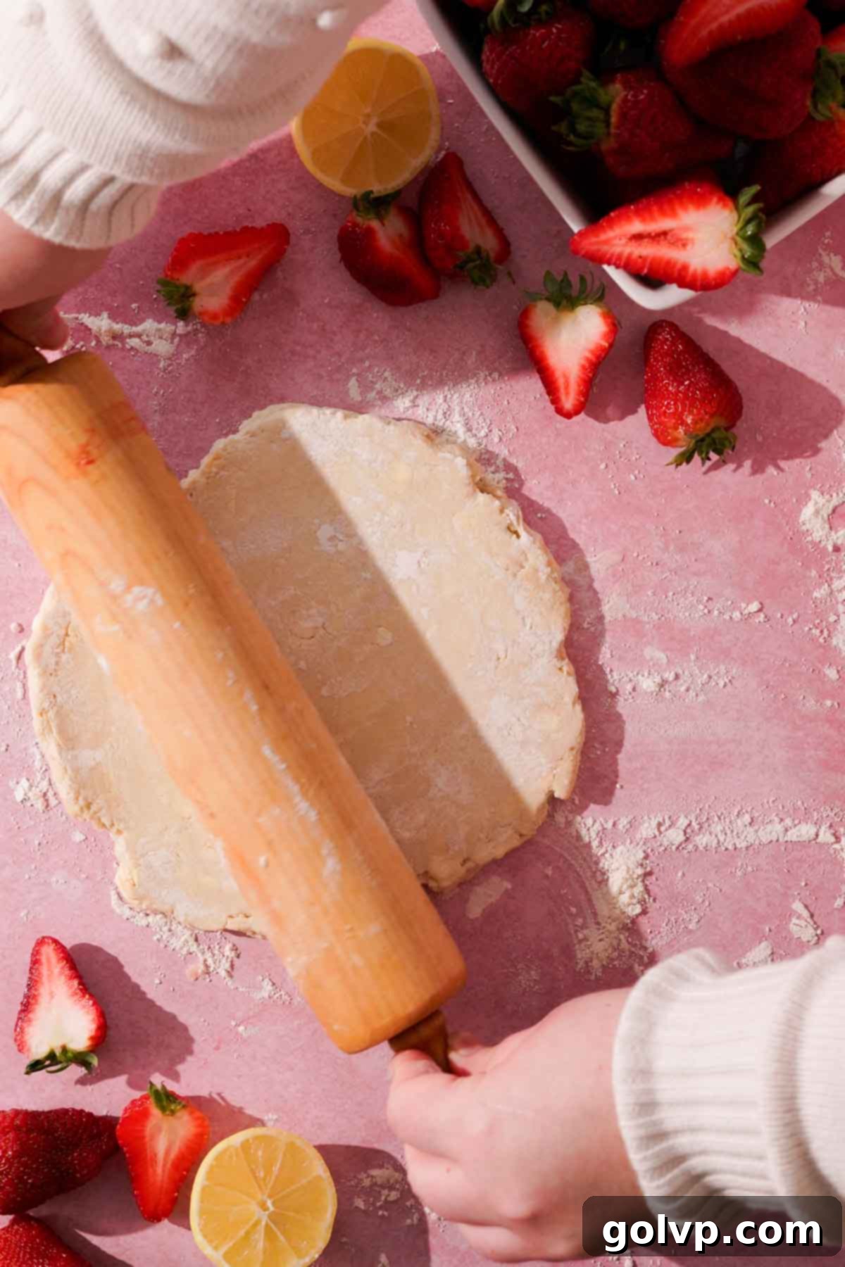 A wooden rolling pin flattening a round disc of galette dough on a pink surface.
