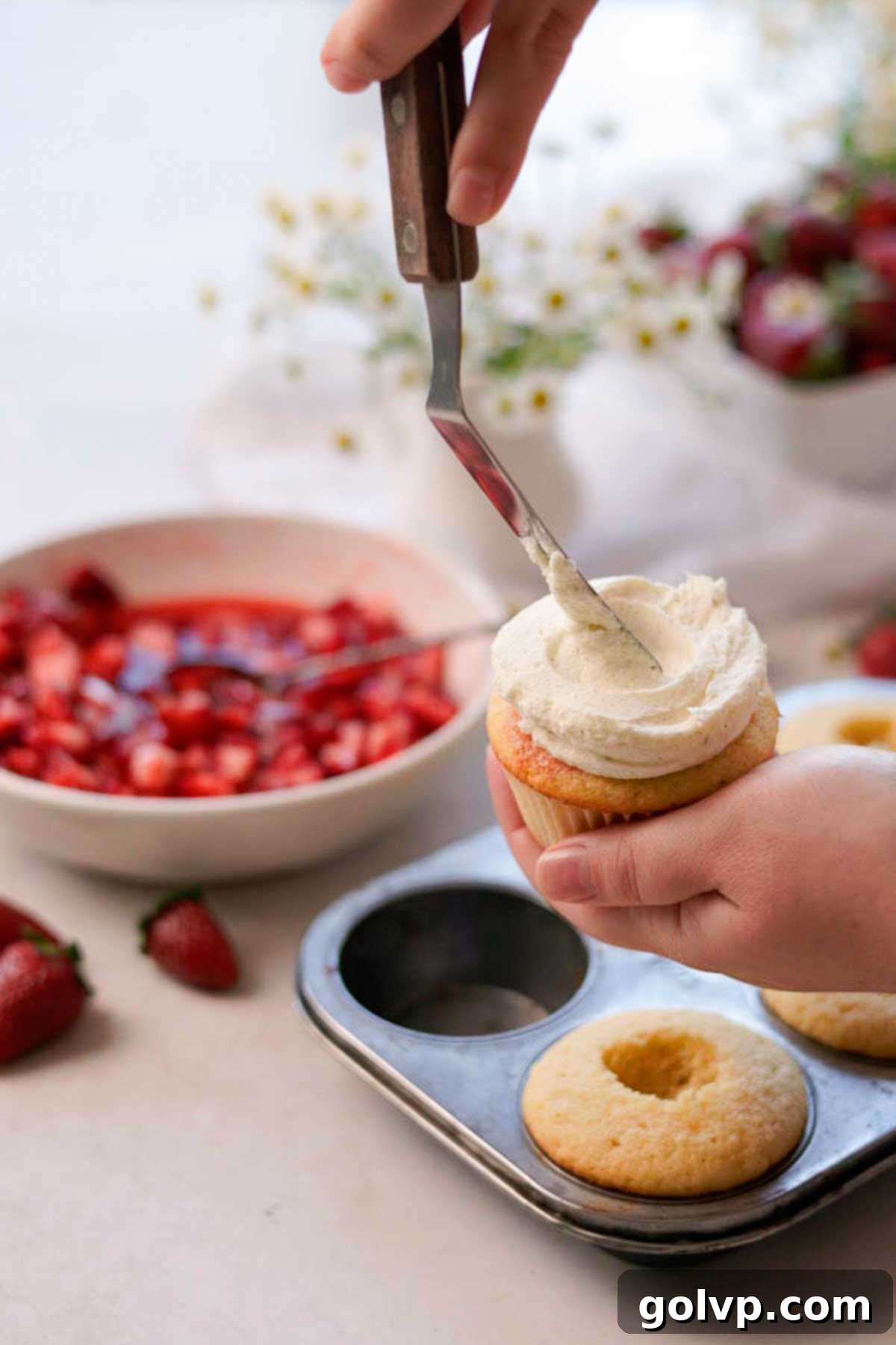 White chocolate mousse frosting being skillfully applied to a strawberry-filled cupcake using a metal spatula, creating a smooth, elegant swirl.