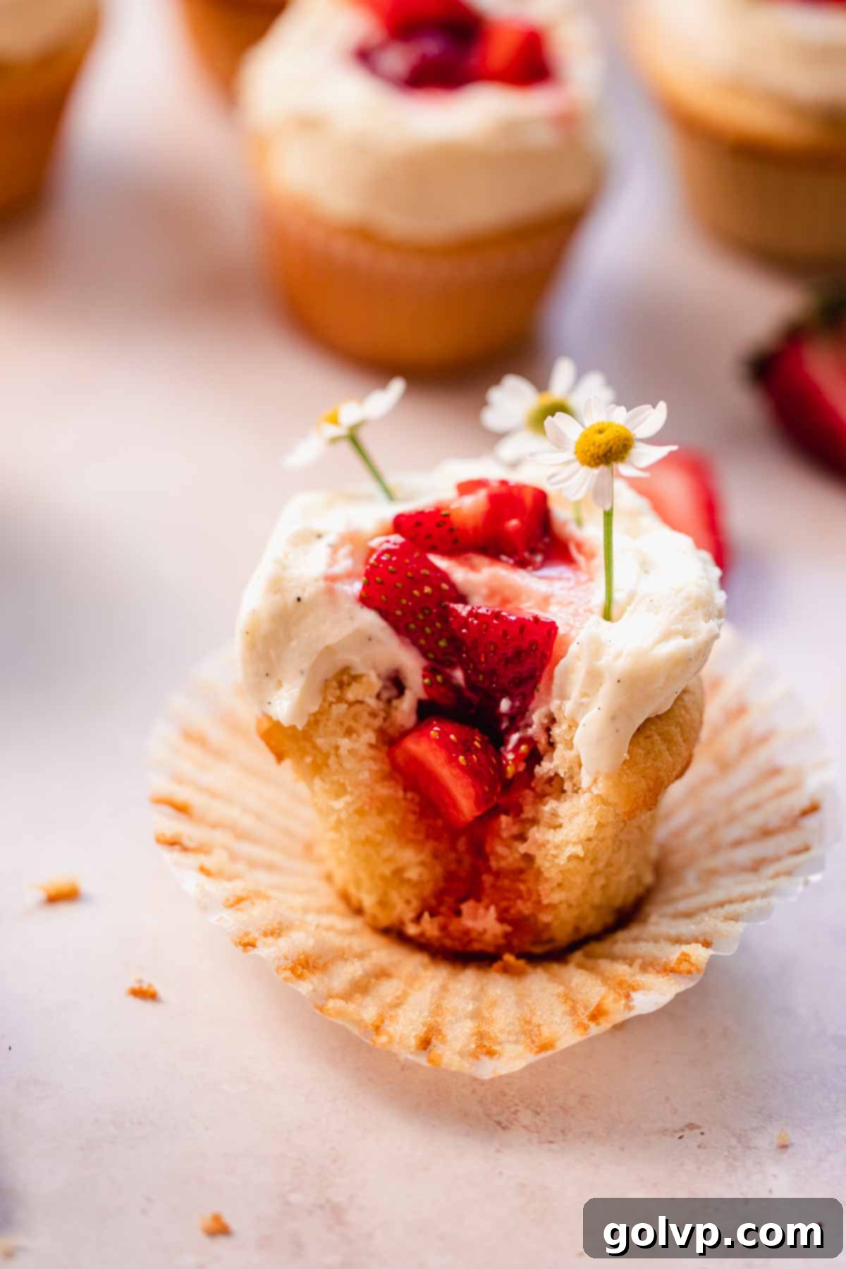 A close-up view of a bitten white chocolate strawberry cupcake, showcasing the juicy strawberry filling and elegant white chocolate mousse frosting with floral garnish.