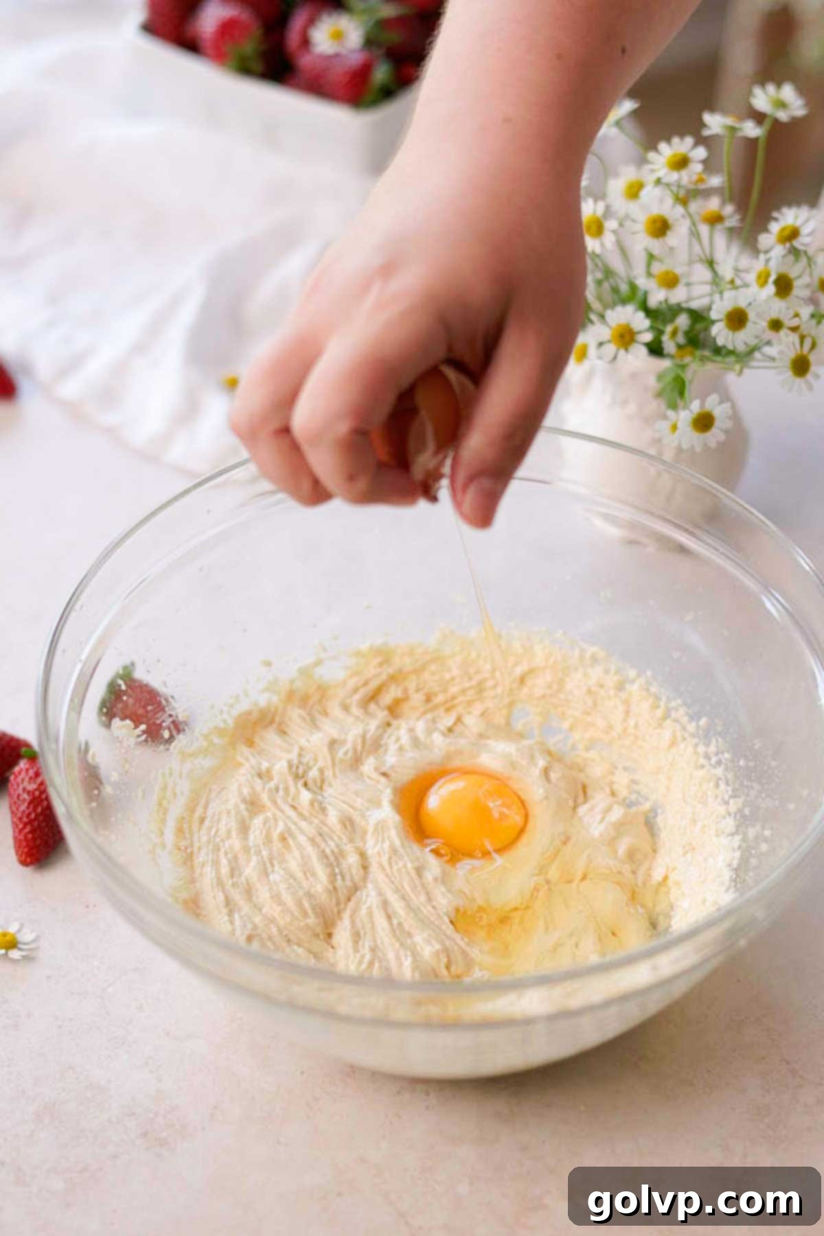 A large egg being cracked and added to the creamy cupcake batter in a glass bowl, ready for mixing.