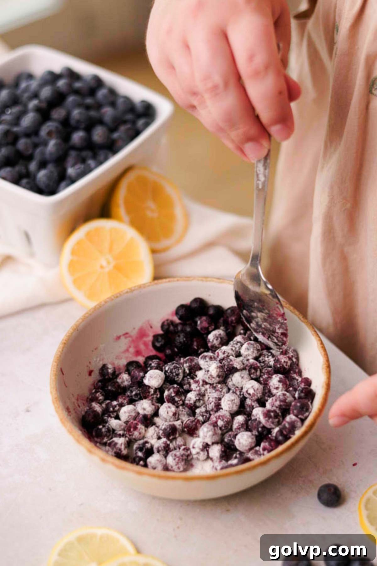 Frozen blueberries being coated with a tablespoon of flour in a bowl.