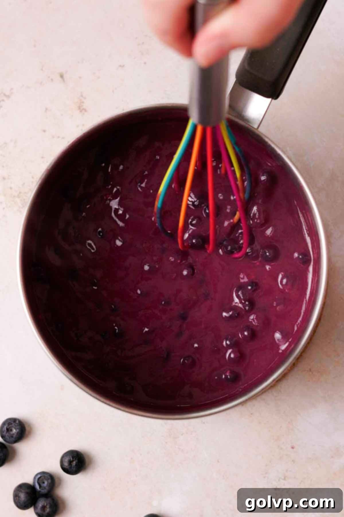 A silicone whisk stirring cooked blueberry curd in a saucepan, showing its thickening consistency.