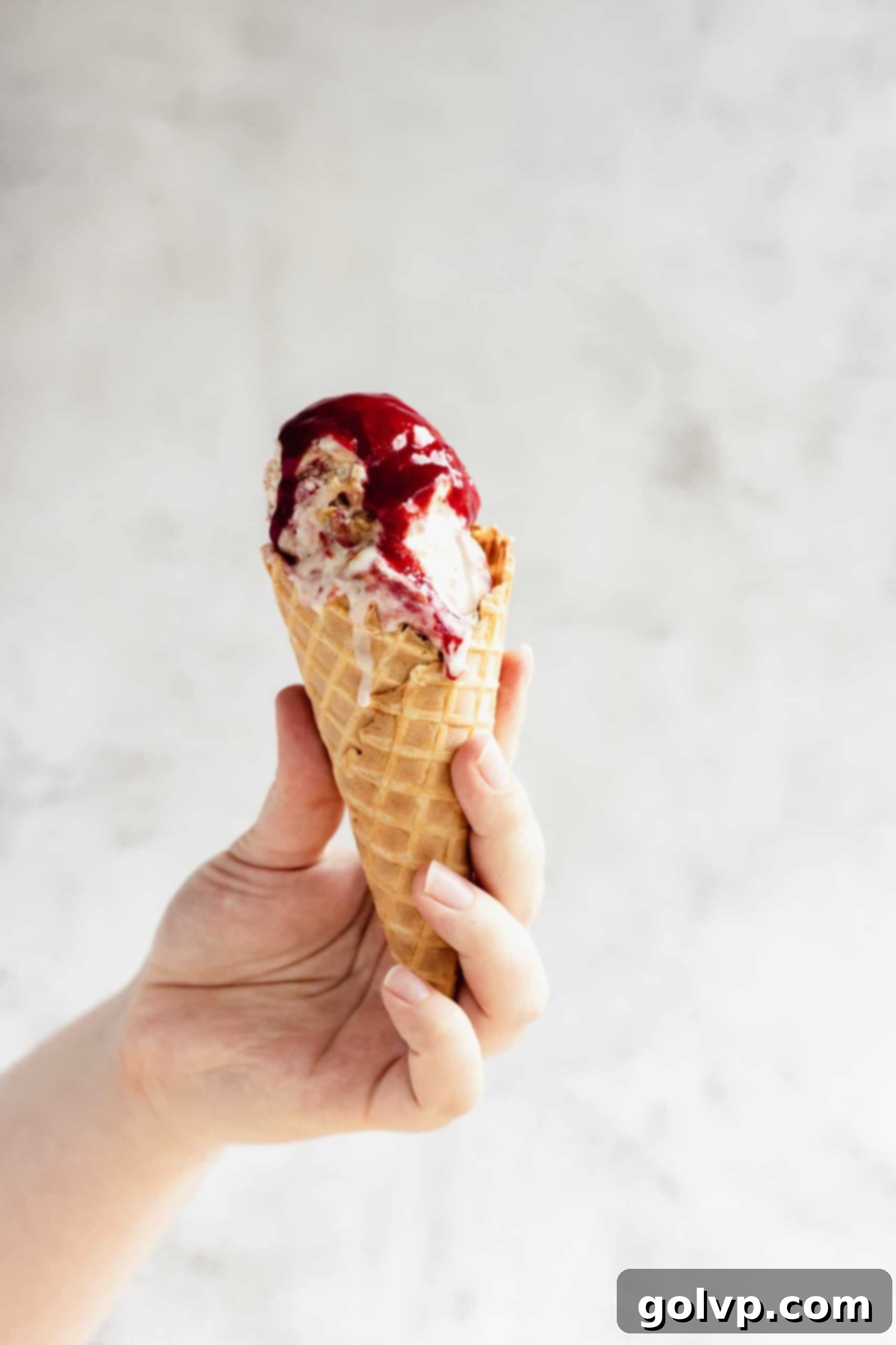 Person holding an ice cream cone topped with raspberry sauce and a hint of graham crumbs