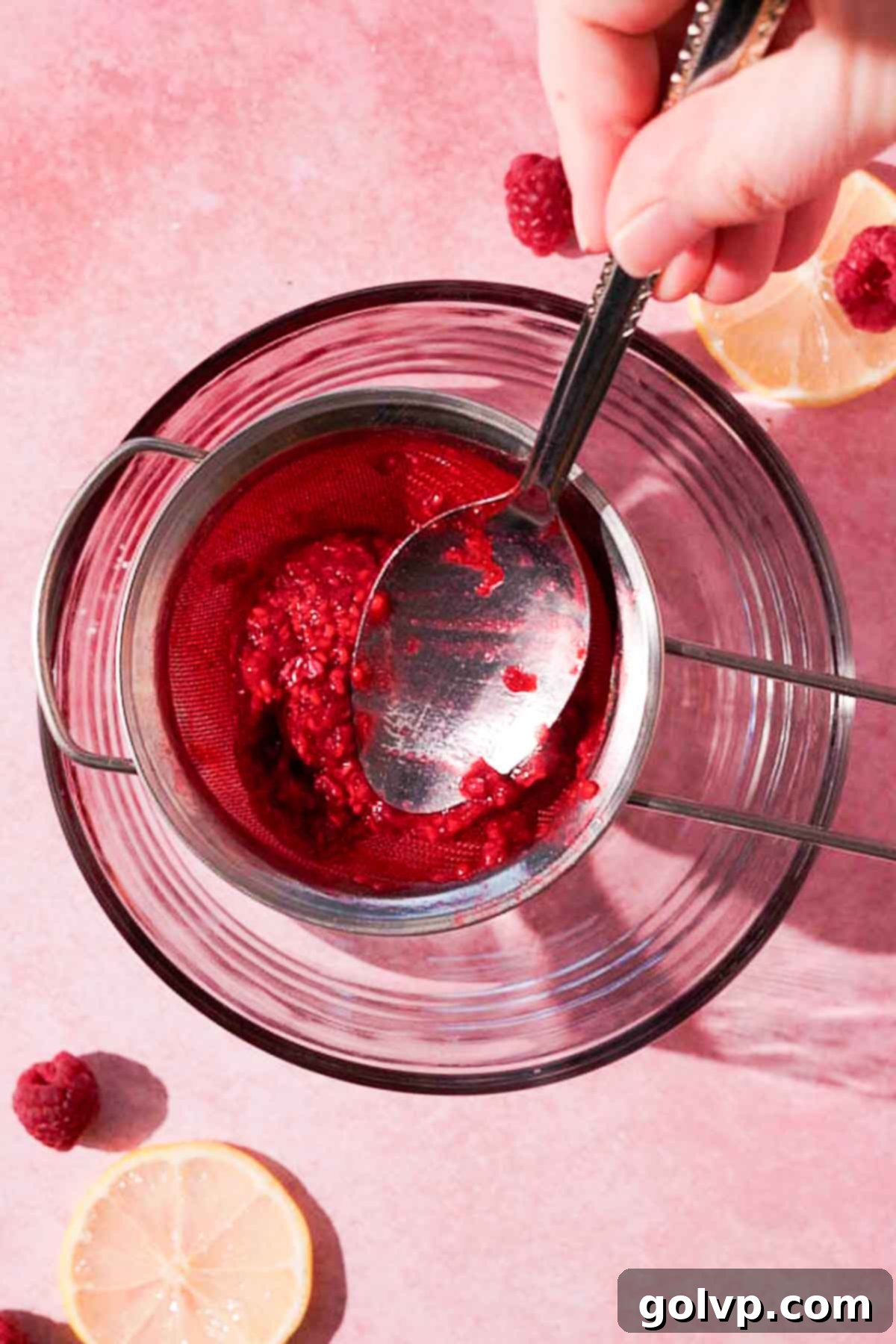 pressing raspberries through a metal sieve into a glass bowl