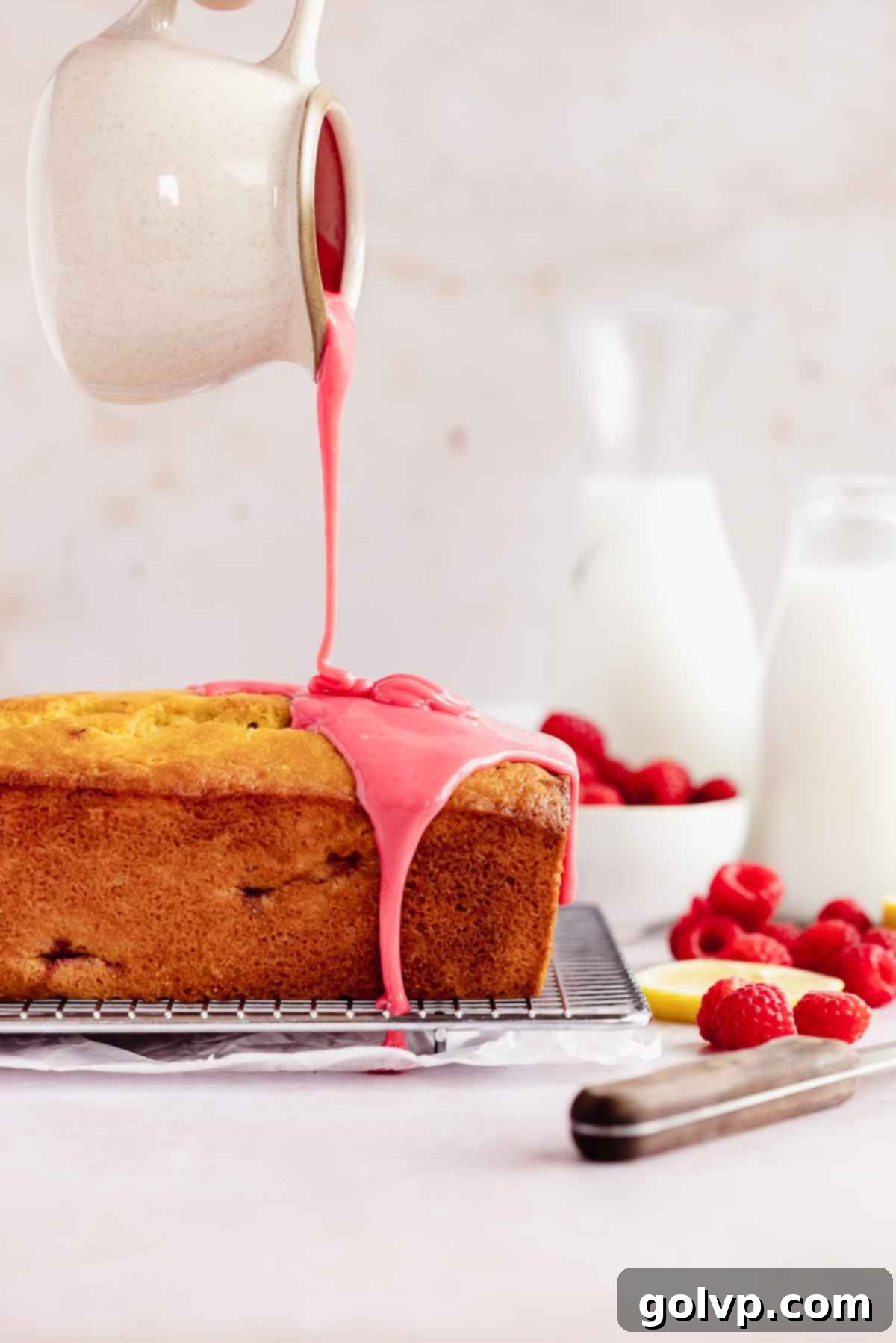 pouring pink glaze from a spout onto a baked loaf cake on cooling rack