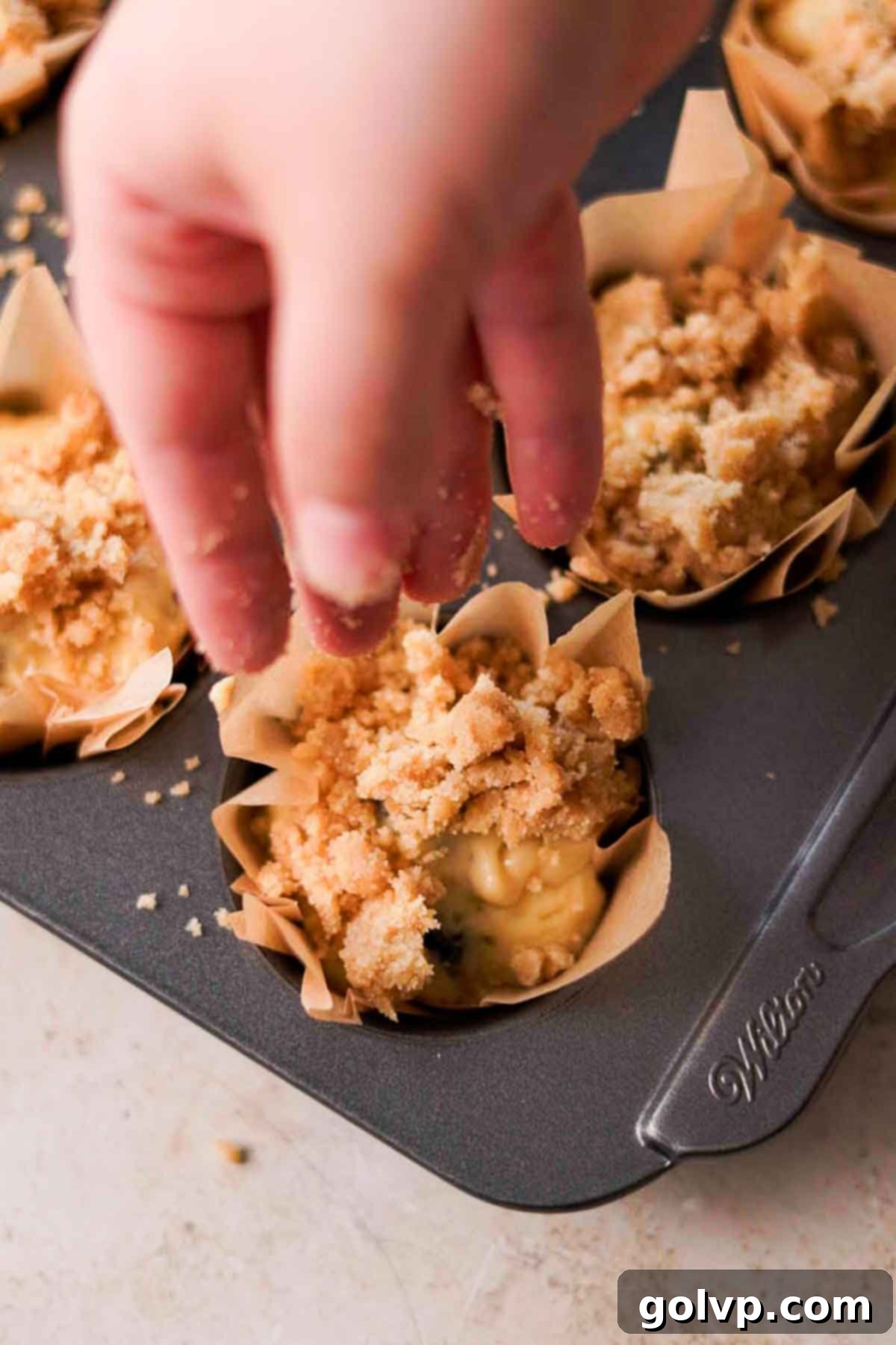Sprinkling a generous amount of crumble topping over muffin batter filled in liners.
