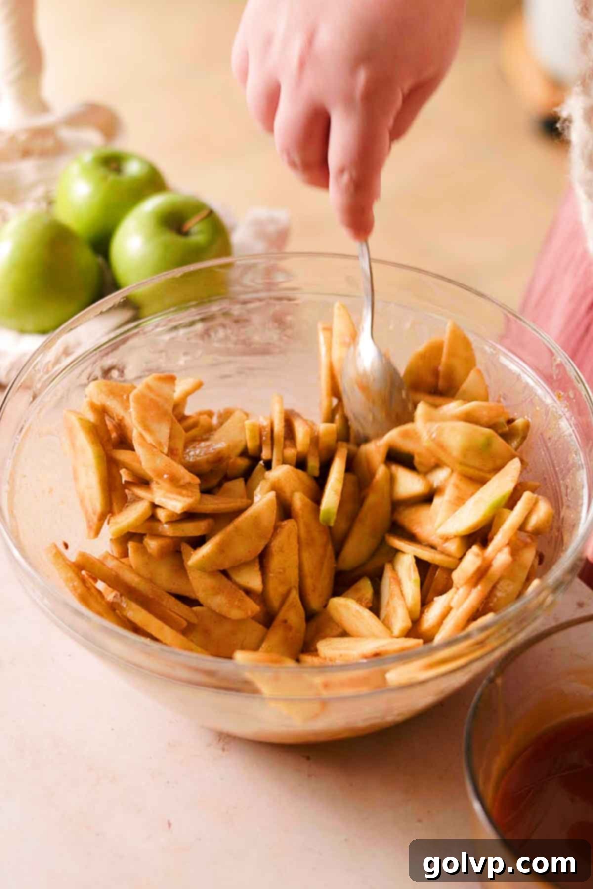 Stirring a large glass bowl of caramel apple pie filling ingredients, ensuring all apple slices are evenly coated.