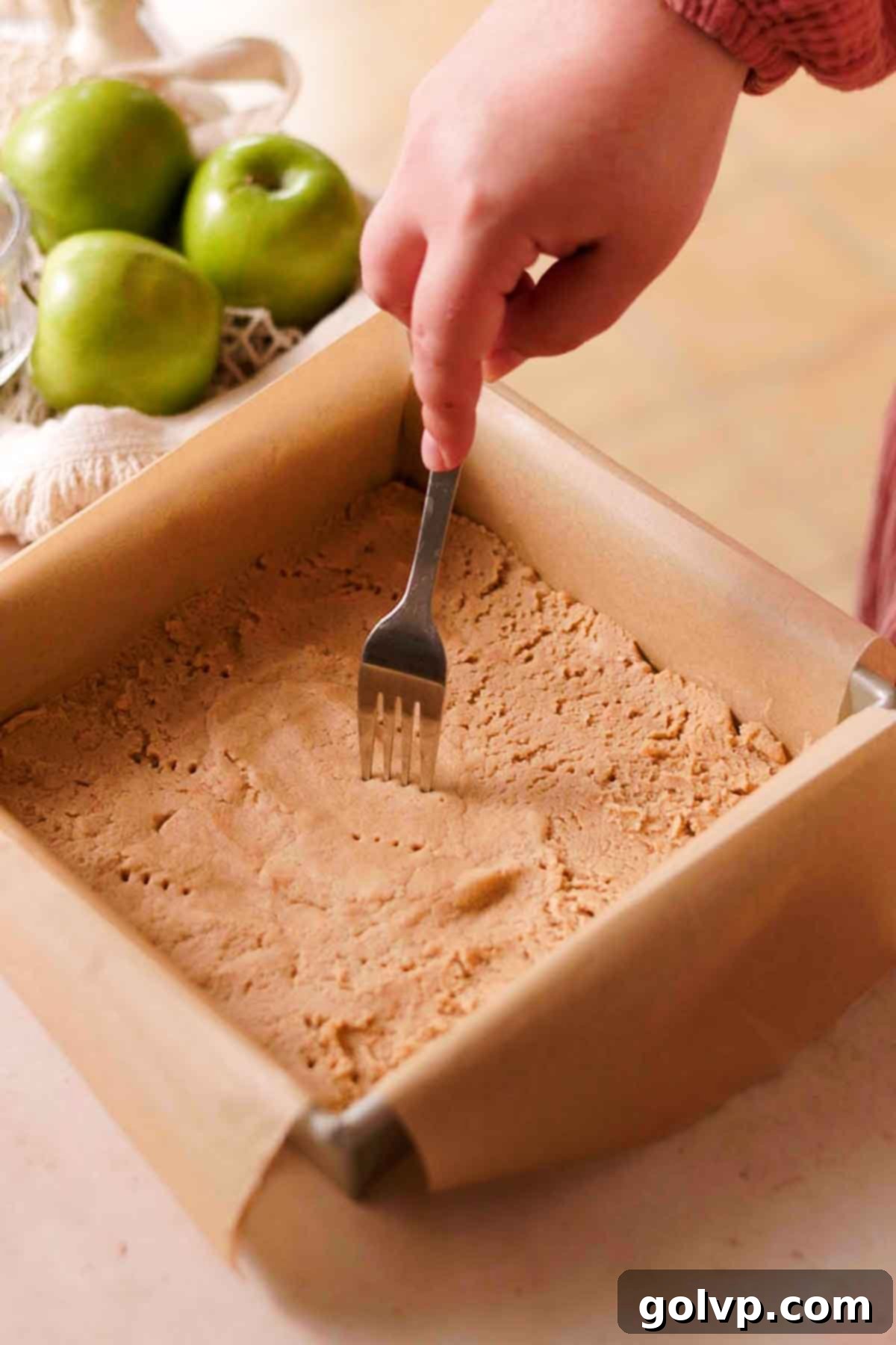 Pricking the shortbread crust evenly with a fork in a square baking pan before its initial bake.