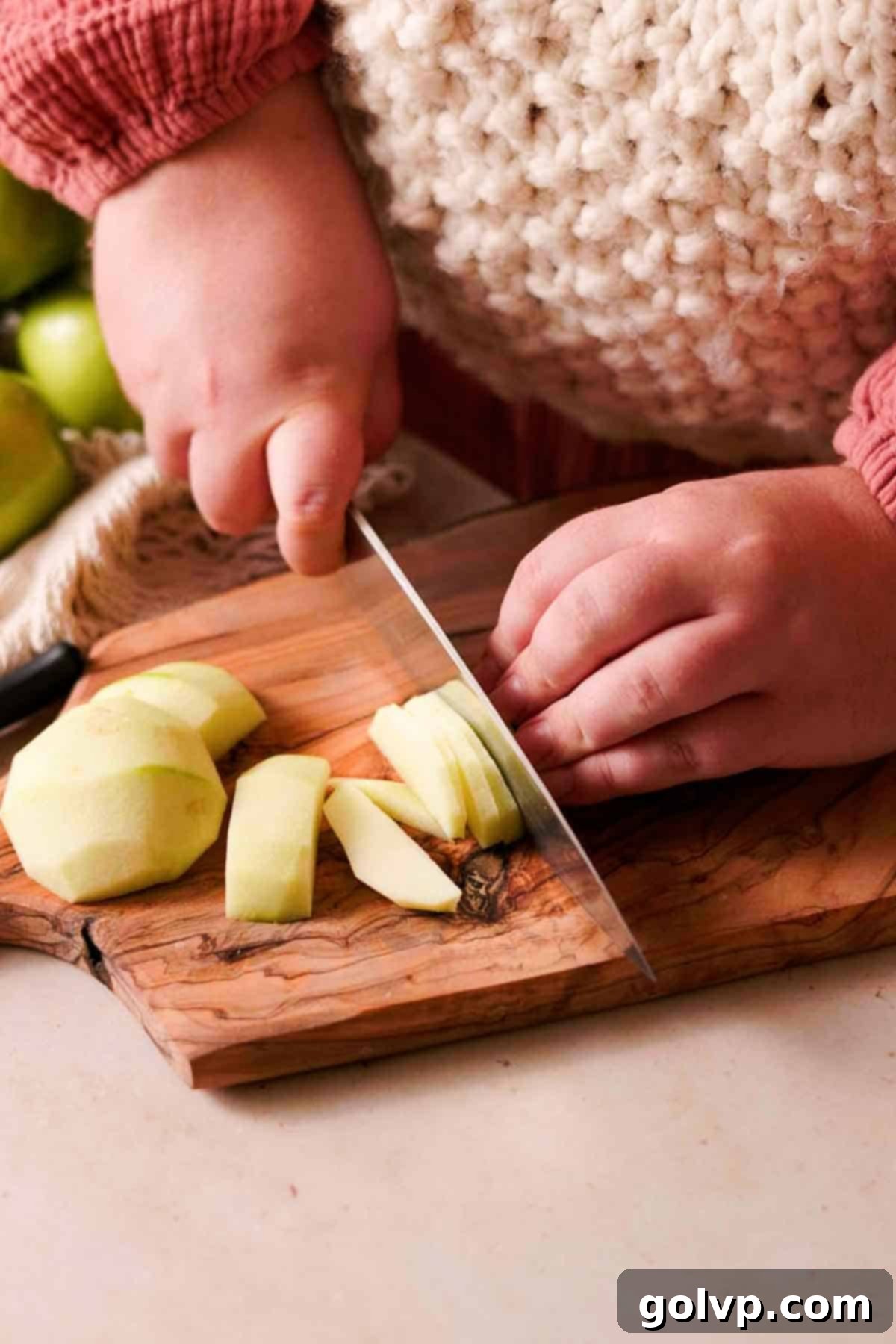 Slicing peeled Granny Smith apples into thin, even pieces on a wooden cutting board with a large knife.