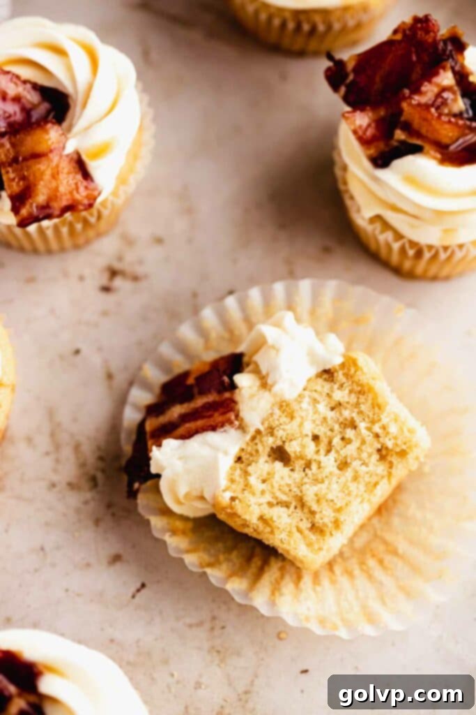 A delicious maple bacon cupcake cut in half, revealing its moist crumb and the rich filling, topped with creamy frosting and candied bacon pieces.