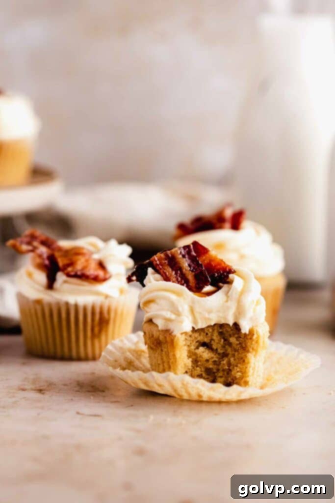 A delicious maple bacon cupcake with a bite taken out, showing the moist cake, creamy frosting, and crispy candied bacon on top.